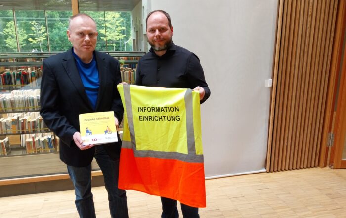 Zwei Männer stehen drinnen vor Bücherregalen; der eine hält eine Broschüre in der Hand, der andere eine gelb-orangefarbene Sicherheitsweste mit der Aufschrift "INFORMATION EINRICHTUNG", die auf Schutzmaßnahmen für Katastrophenfälle hinweist. at Hochschule Coburg