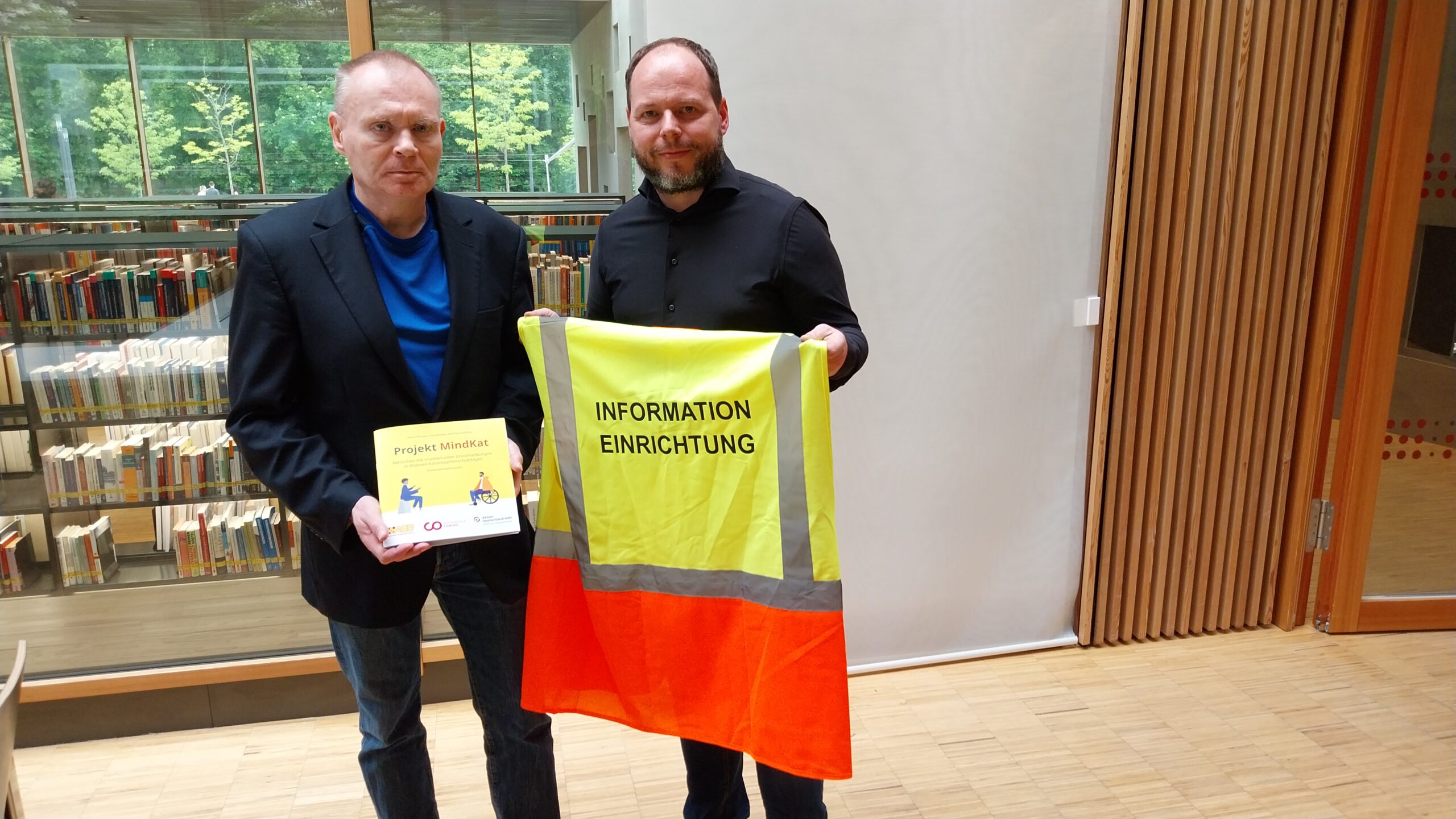 Zwei Männer stehen drinnen vor Bücherregalen; der eine hält eine Broschüre in der Hand, der andere eine gelb-orangefarbene Sicherheitsweste mit der Aufschrift "INFORMATION EINRICHTUNG", die auf Schutzmaßnahmen für Katastrophenfälle hinweist. at Hochschule Coburg