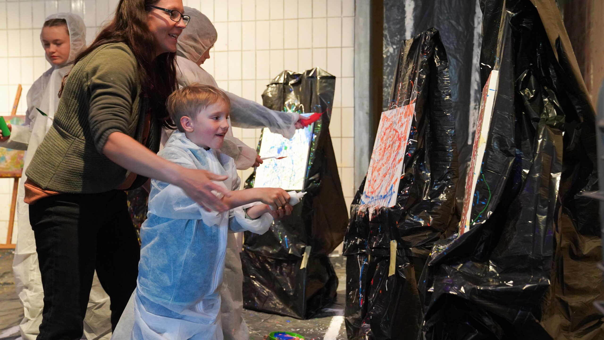 Eine Frau und ein lächelndes Kind in Schutzanzügen bemalen bei der Langen Nacht der Wissenschaften Leinwände, die mit Plastik bedeckt sind, und schaffen so eine lebendige Resonanz. Zwei weitere Personen stehen in der Nähe, die ebenfalls für diese kreative, chaotische Kunstumgebung gekleidet sind. at Hochschule Coburg