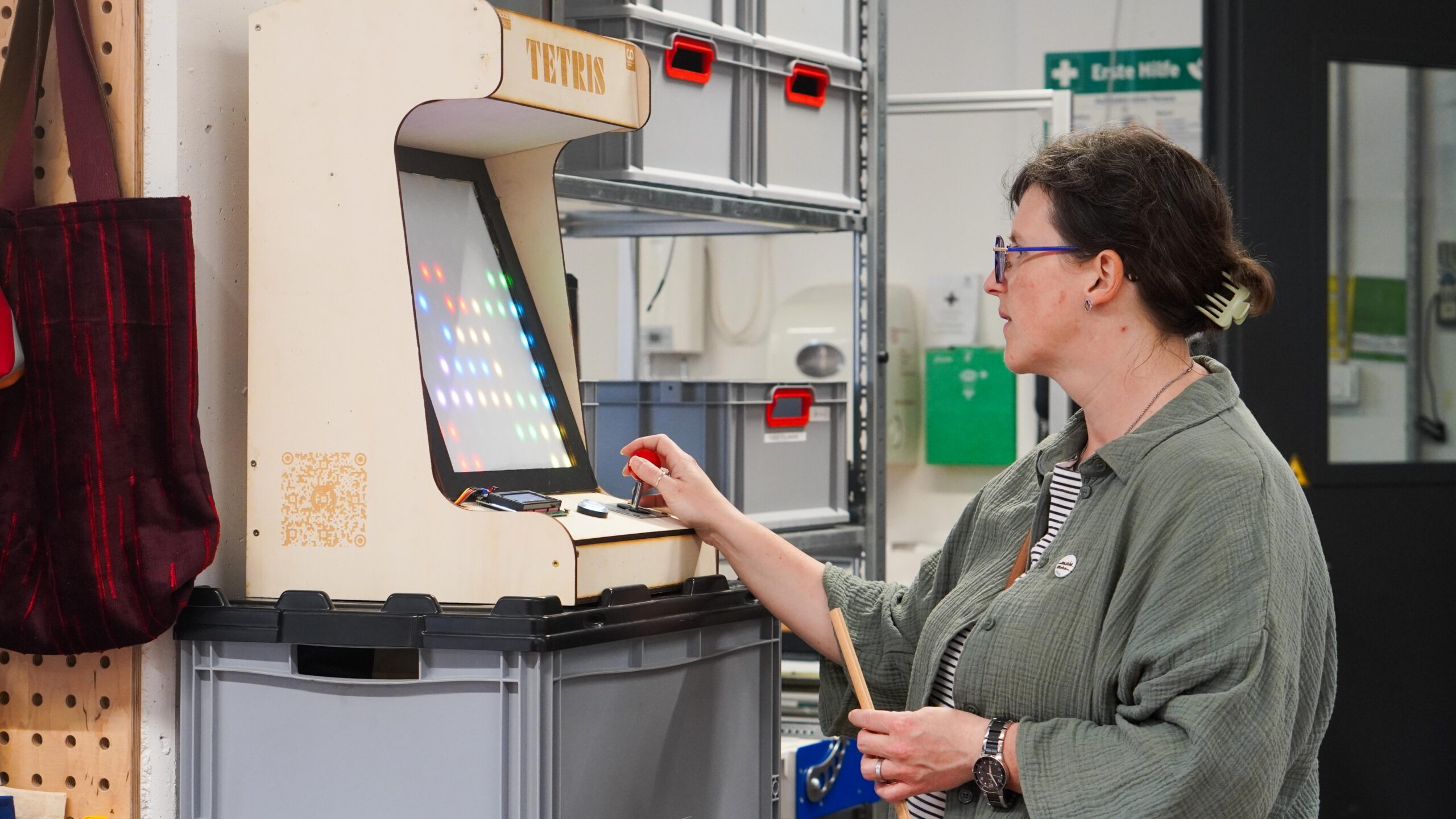 Eine Person mit Brille und grünem Hemd spielt während der Langen Nacht der Wissenschaften in einer Werkstatt ein Tetris-Arcade-Spiel auf einem Holzschrank, umgeben von Regalen und Werkzeugen - eine perfekte Mischung aus Spielen und Wissenschaften. at Hochschule Coburg
