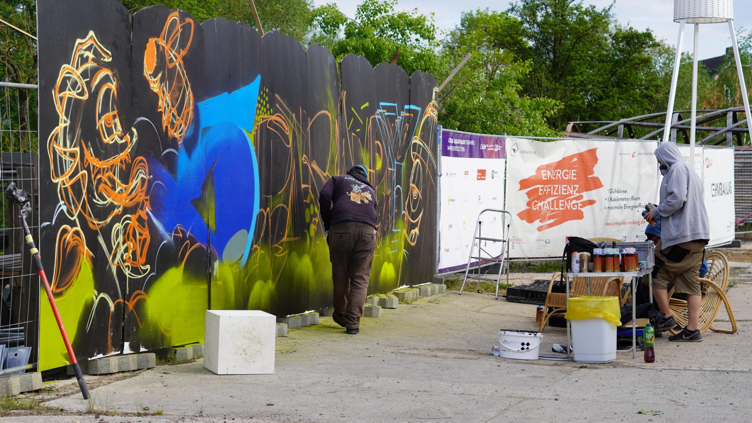 Zwei Personen in Kapuzenpullovern sprühen während der Langen Nacht der Wissenschaften bunte Graffiti-Kunst auf einen schwarzen Holzzaun im Freien, während Farbdosen und Zubehör auf dem Boden liegen und im Hintergrund ein Transparent mit der Aufforderung zum Mitmachen zu sehen ist. at Hochschule Coburg
