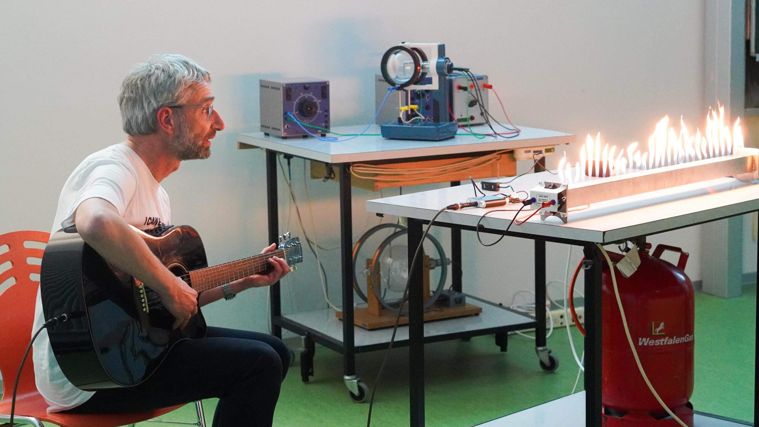 Ein Mann spielt während der Langen Nacht der Wissenschaften in einem Klassenzimmer auf einer Akustikgitarre und beobachtet dabei ein Flammenrohr-Experiment. Die Flammen erzeugen mit den Schallwellen eine enorme Resonanz, die inmitten von wissenschaftlichen Geräten und einer roten Gasflasche auffällige Wellenmuster bildet. at Hochschule Coburg