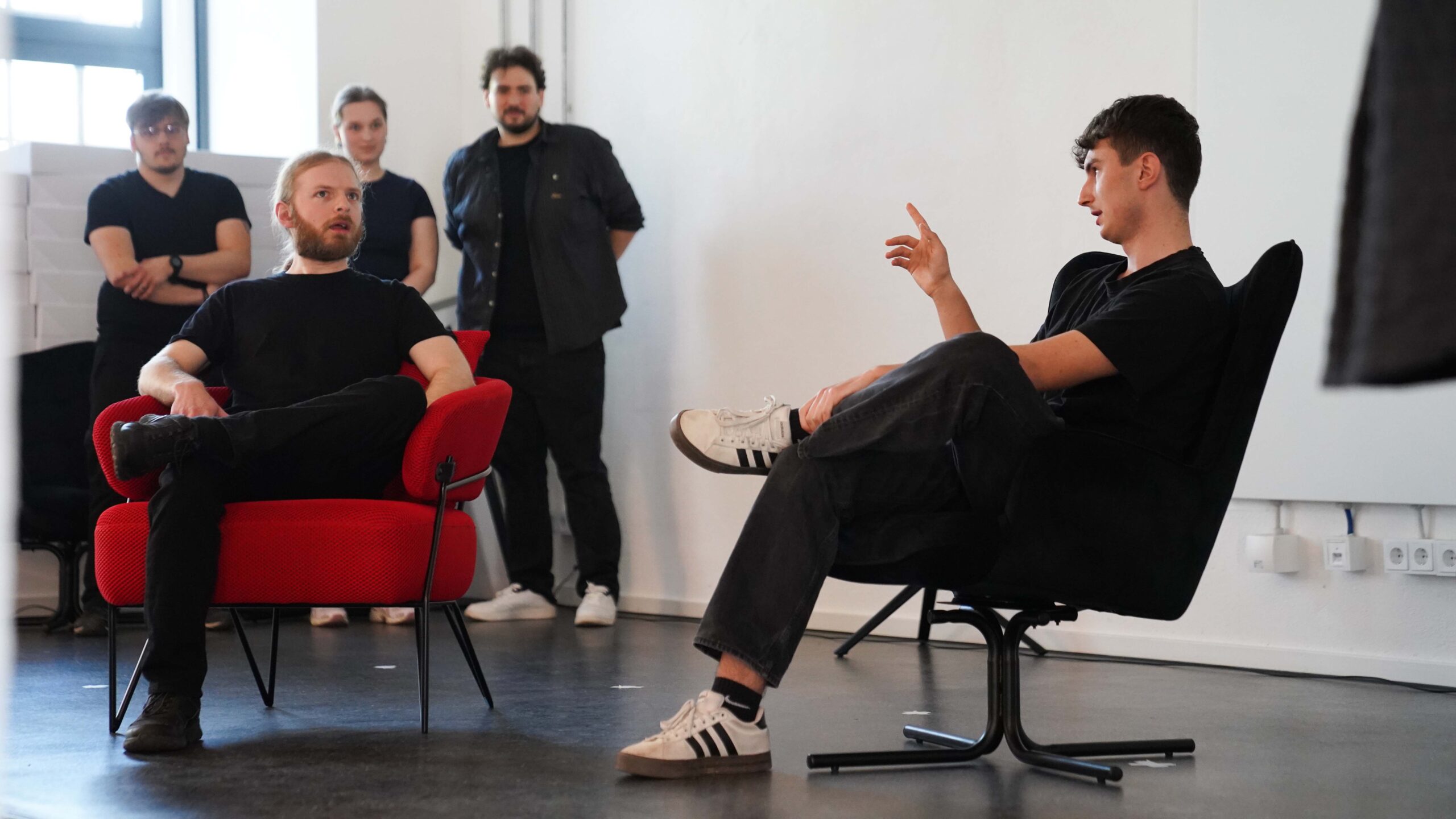 Zwei Männer sitzen sich auf rot-schwarzen Stühlen gegenüber und unterhalten sich über Talente und Unternehmen, während im Hintergrund eine Gruppe von Menschen steht und sie in einem hellen, modernen Raum beobachtet. at Hochschule Coburg