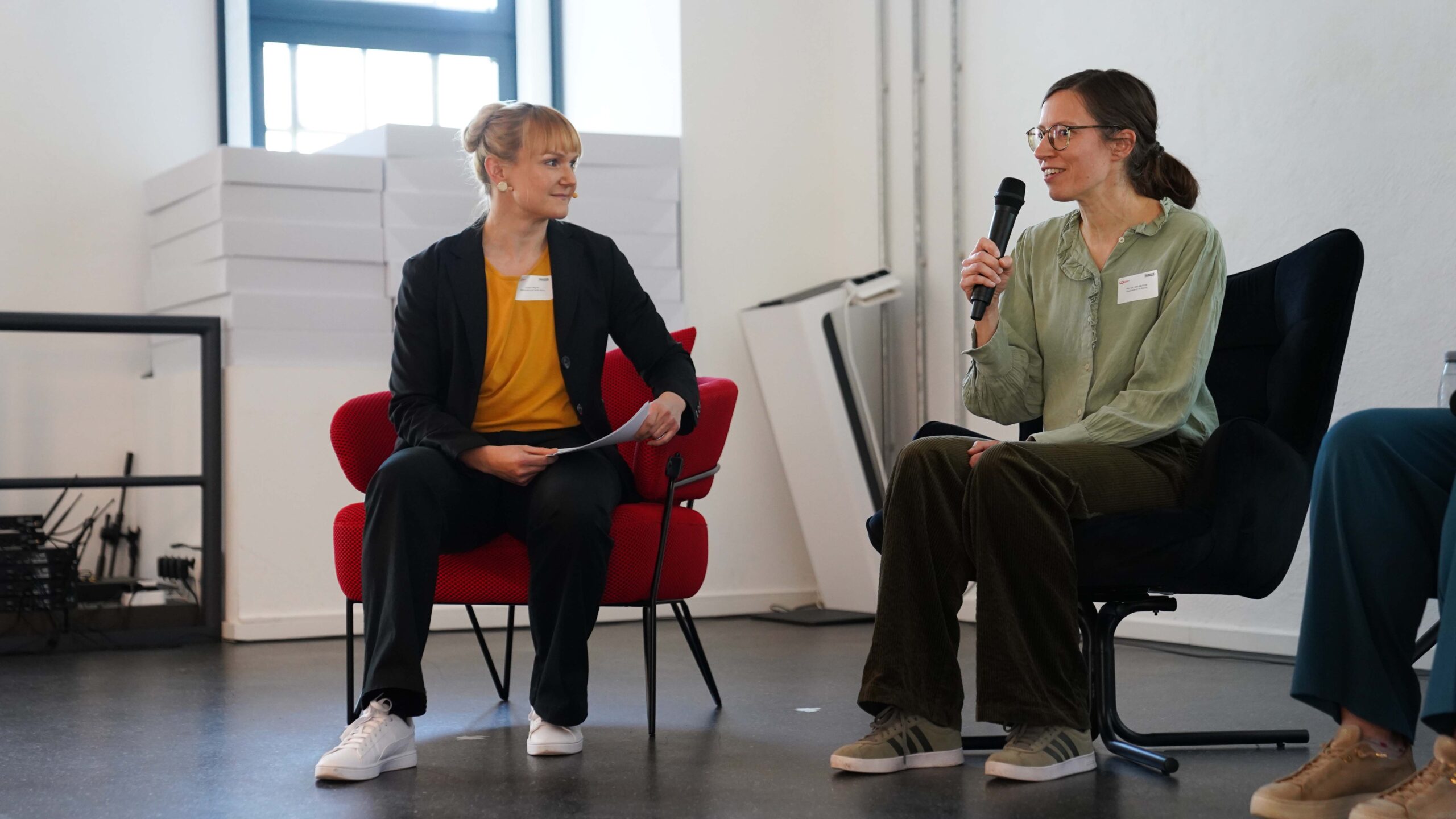 Zwei Frauen sitzen und unterhalten sich, eine hält ein Mikrofon, die andere hört zu und lächelt. In einem modernen, hell erleuchteten Raum diskutieren sie darüber, wie die Investition in Talente, zum Beispiel durch das Deutschlandstipendium, Mehrwert für Unternehmen schafft. at Hochschule Coburg