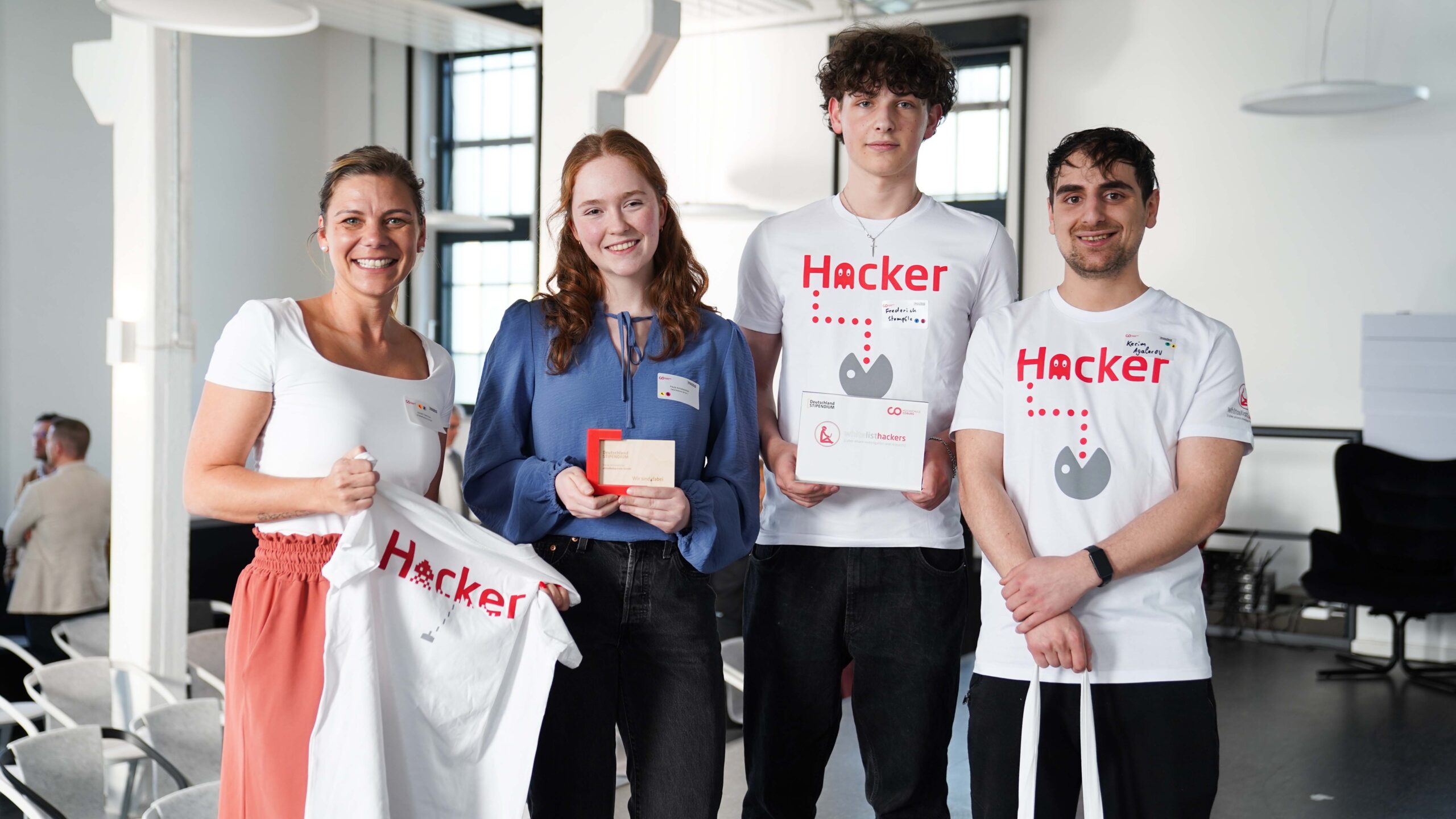 Vier Personen stehen in einem Haus und lächeln in die Kamera. Zwei tragen "Hacker"-Shirts, die anderen halten Geschenke und ein T-Shirt. Sie scheinen auf einer Veranstaltung zu sein, auf der Talente oder ein Deutschlandstipendium in Zusammenarbeit mit Unternehmen gefeiert werden. at Hochschule Coburg