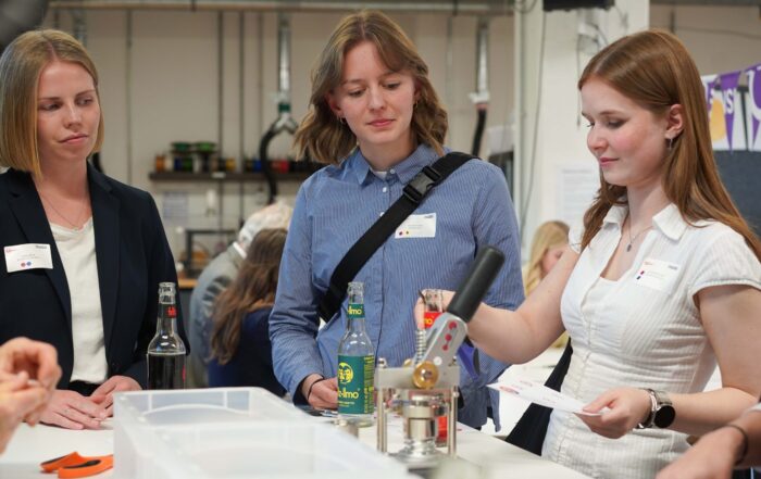 Drei Frauen stehen bei einer Veranstaltung um einen Tisch herum. Eine demonstriert die Verwendung einer Maschine, während die anderen zusehen und Talente präsentieren. Auf dem Tisch stehen Flaschen und Materialien, und im Hintergrund arbeiten Menschen. at Hochschule Coburg