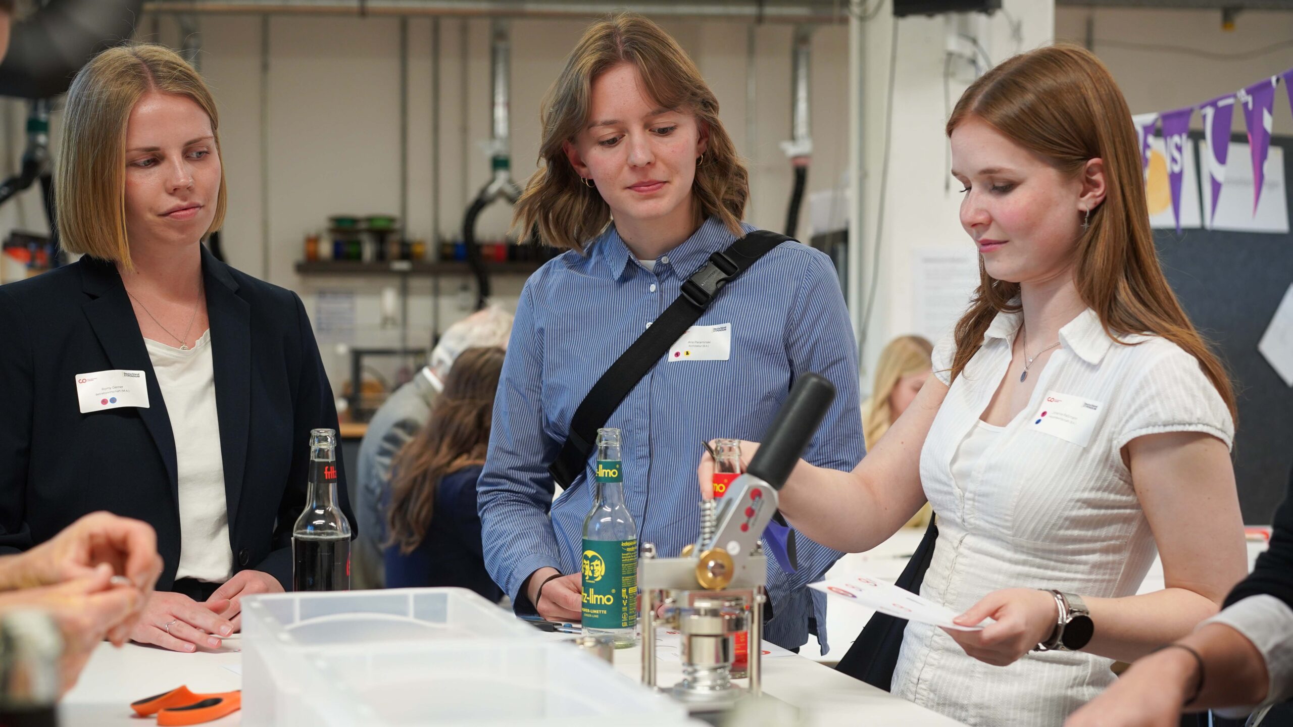 Drei Frauen stehen bei einer Veranstaltung um einen Tisch herum. Eine demonstriert die Verwendung einer Maschine, während die anderen zusehen und Talente präsentieren. Auf dem Tisch stehen Flaschen und Materialien, und im Hintergrund arbeiten Menschen. at Hochschule Coburg