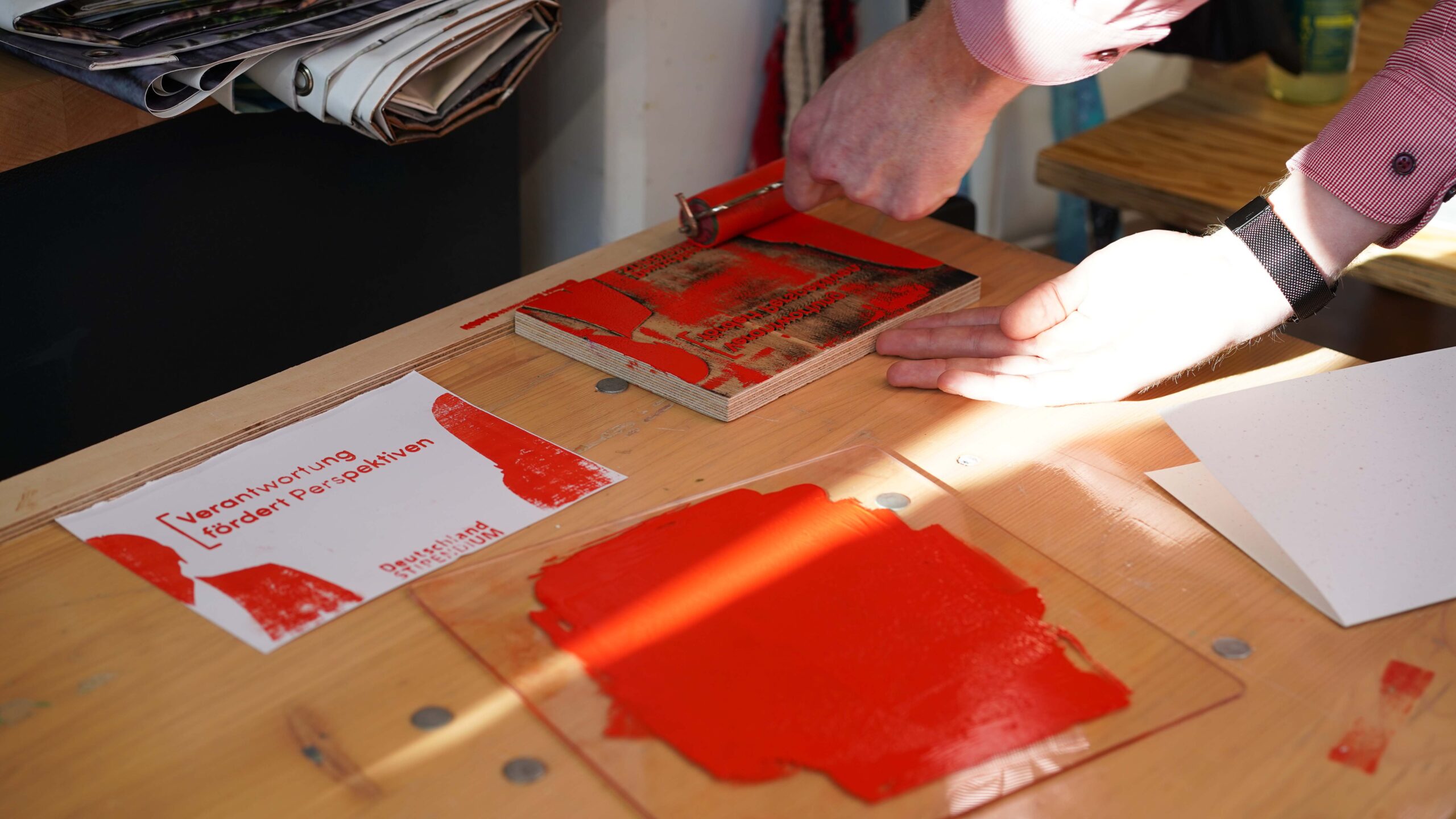 Eine Person rollt rote Tinte auf ein hölzernes Klischee auf einem Tisch und bereitet sich auf die Druckvorbereitung vor. In der Nähe befinden sich eine Glasplatte mit roter Tinte, Papier und eine gedruckte Karte mit einem deutschen Text über Talente und Unternehmen im Deutschlandstipendium-Programm. at Hochschule Coburg