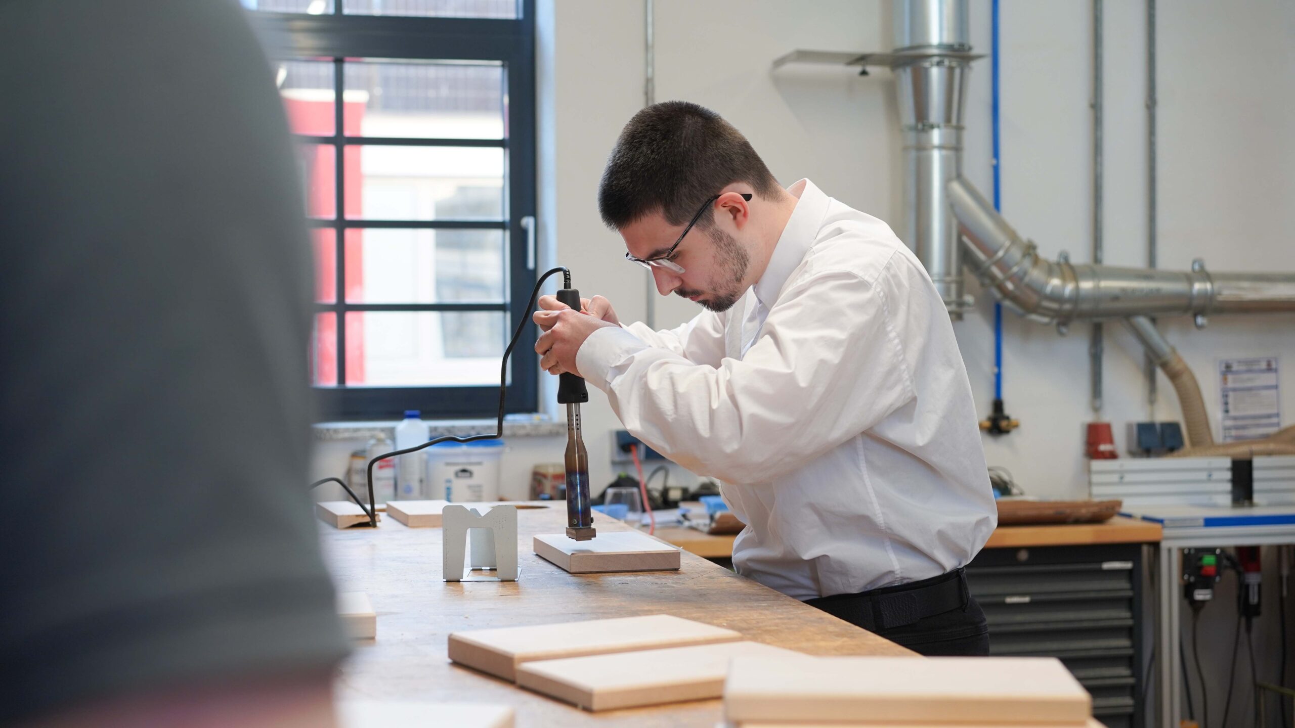 Eine Person im weißen Hemd arbeitet mit einer Bohrmaschine an einem Stück Holz in einer Werkstatt, umgeben von Werkzeugen und Geräten, in die Licht durch ein großes Fenster fällt - der perfekte Ort für die vom Deutschlandstipendium geförderten Talente, um zu kreieren und zu innovieren. at Hochschule Coburg