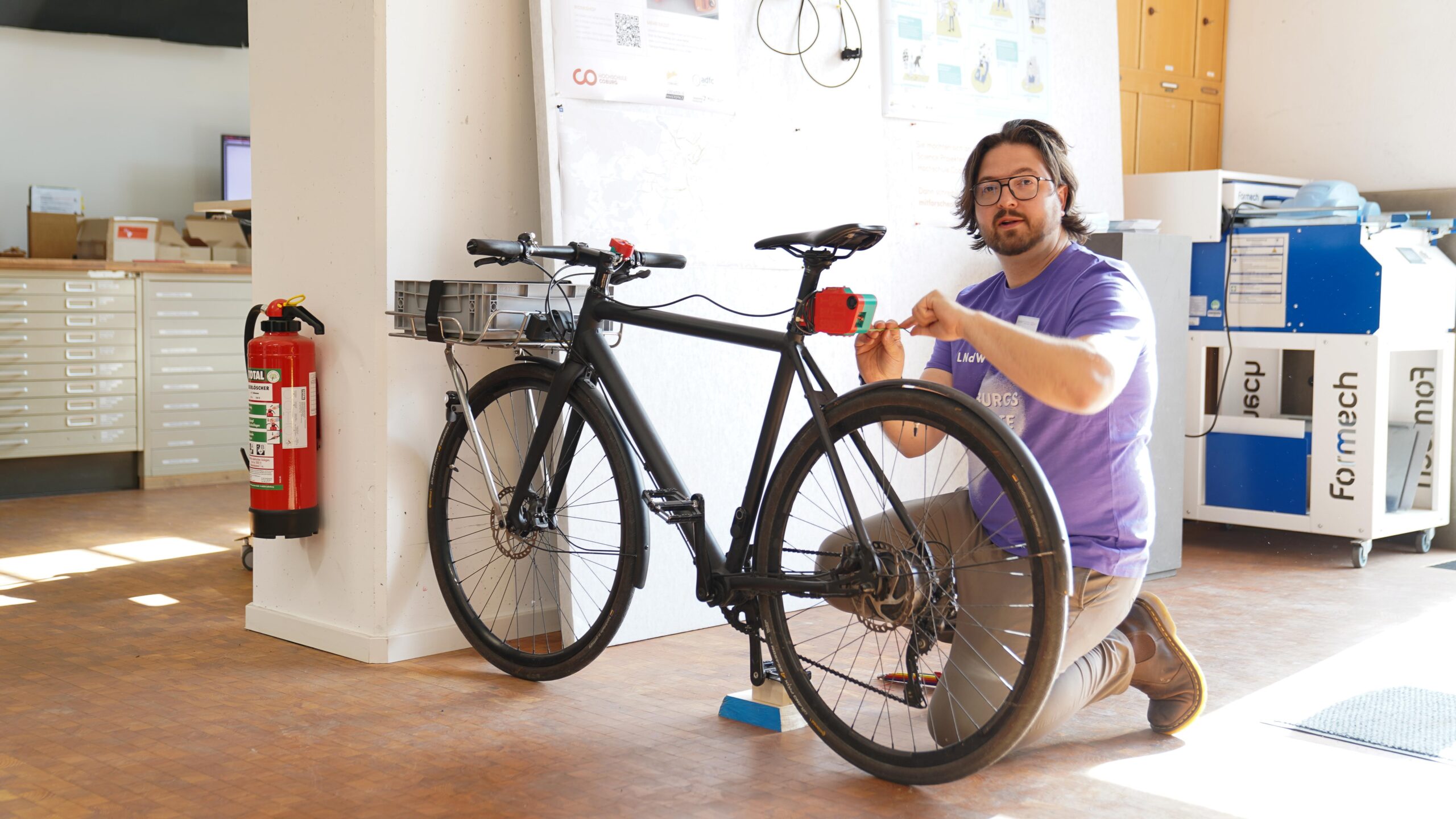 Eine Person mit Brille und lilafarbenem Hemd kniet in einem Innenraum neben einem schwarzen Fahrrad und stellt den Lenker ein. Der Arbeitsbereich, der wahrscheinlich für die Lange Nacht der Wissenschaften eingerichtet wurde, verfügt über Werkzeuge, Schränke und einen Feuerlöscher im Hintergrund. at Hochschule Coburg