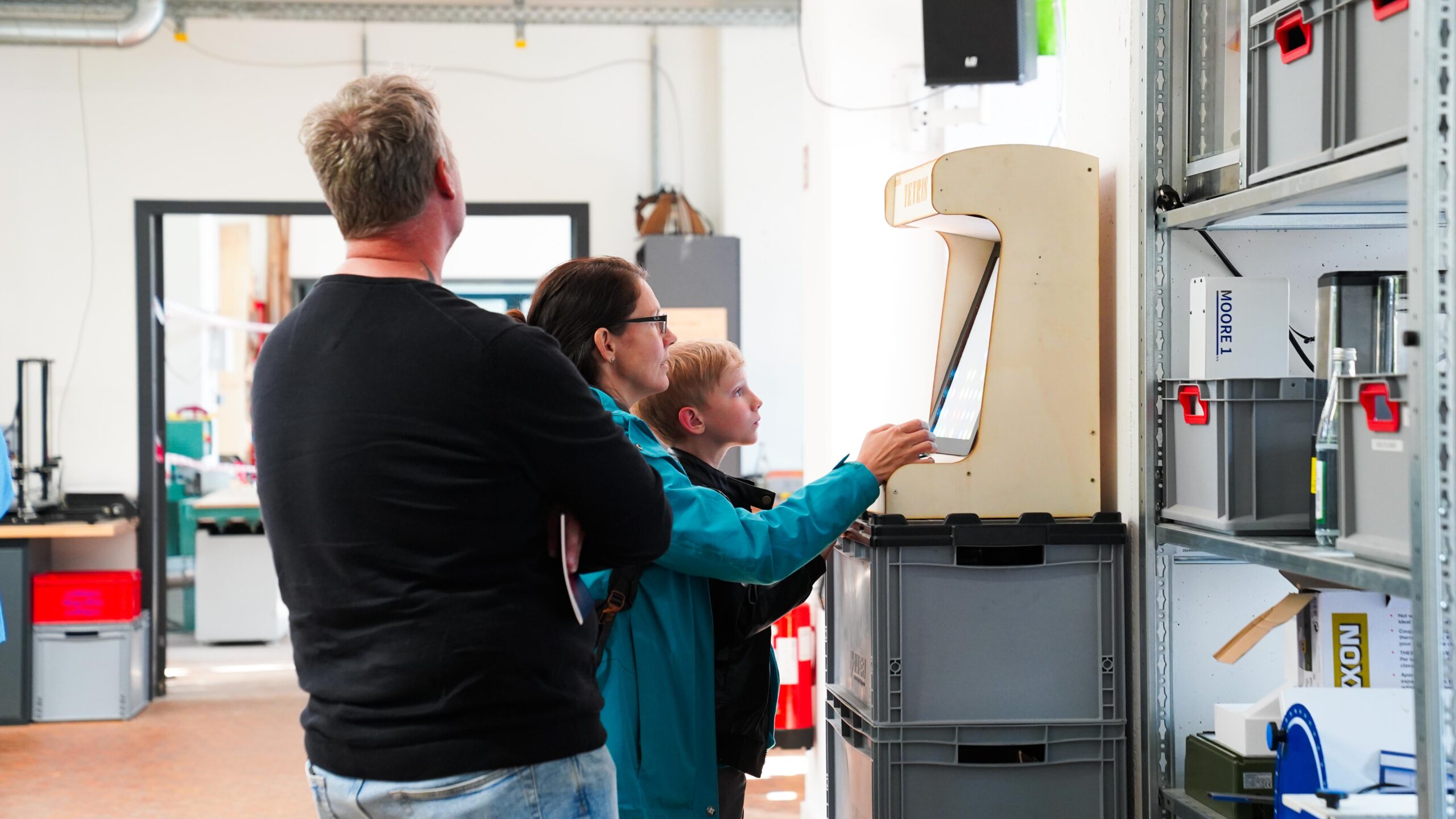 Eine Frau und ein kleiner Junge benutzen einen alten Computer, während ein Mann neben ihnen in einem hellen, modernen Raum der Langen Nacht der Wissenschaften steht, umgeben von Regalen und Geräten, die eine besondere Resonanz von Vergangenheit und Innovation erzeugen. at Hochschule Coburg