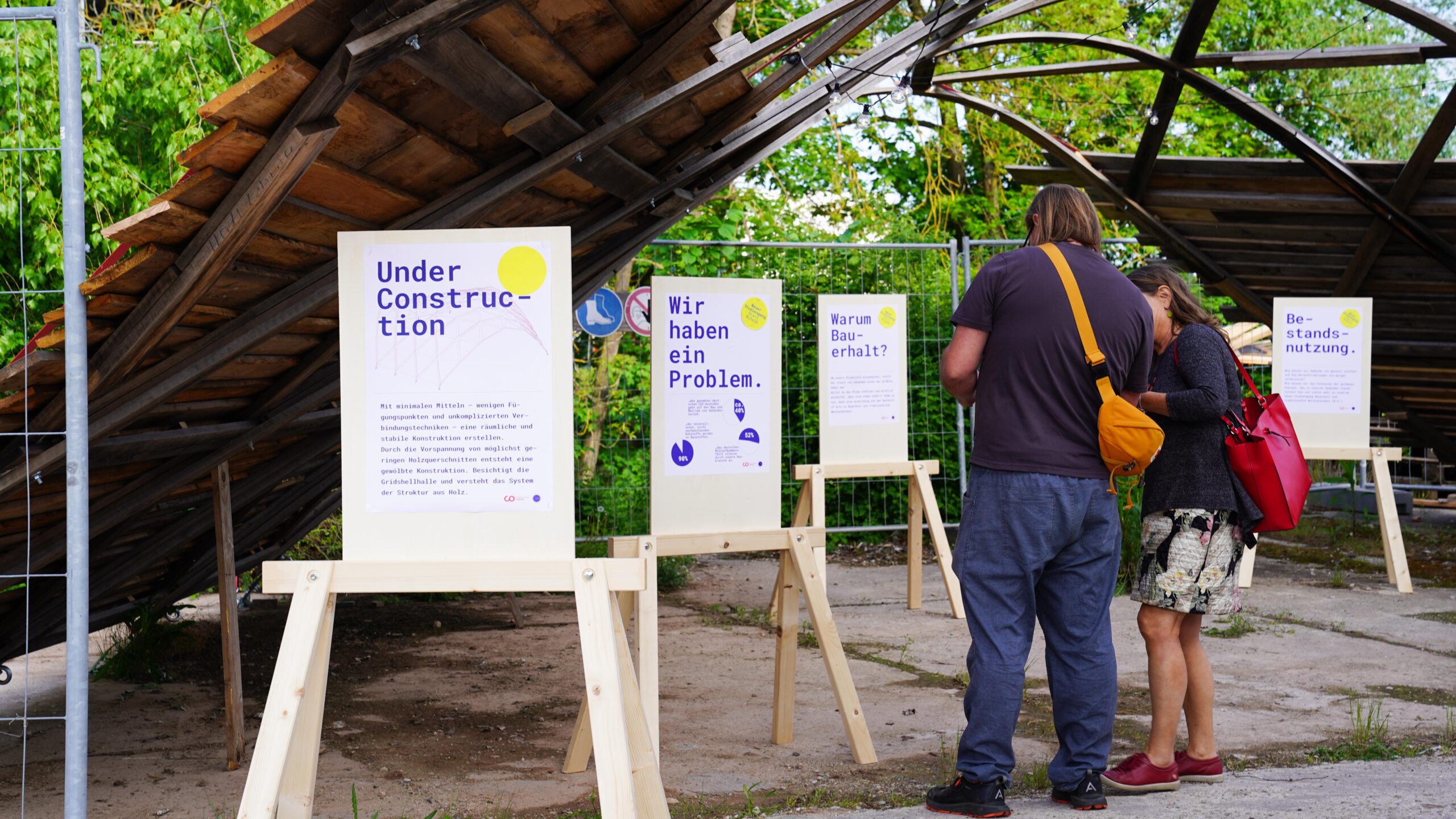 Zwei Personen lesen Informationsplakate auf Holzständern unter einer teilweise eingestürzten Holzkonstruktion, umgeben von Grünpflanzen und einem Metallzaun. Die Plakate, die Teil der Langen Nacht der Wissenschaften sind, bieten Einblicke in deutscher und englischer Sprache. at Hochschule Coburg