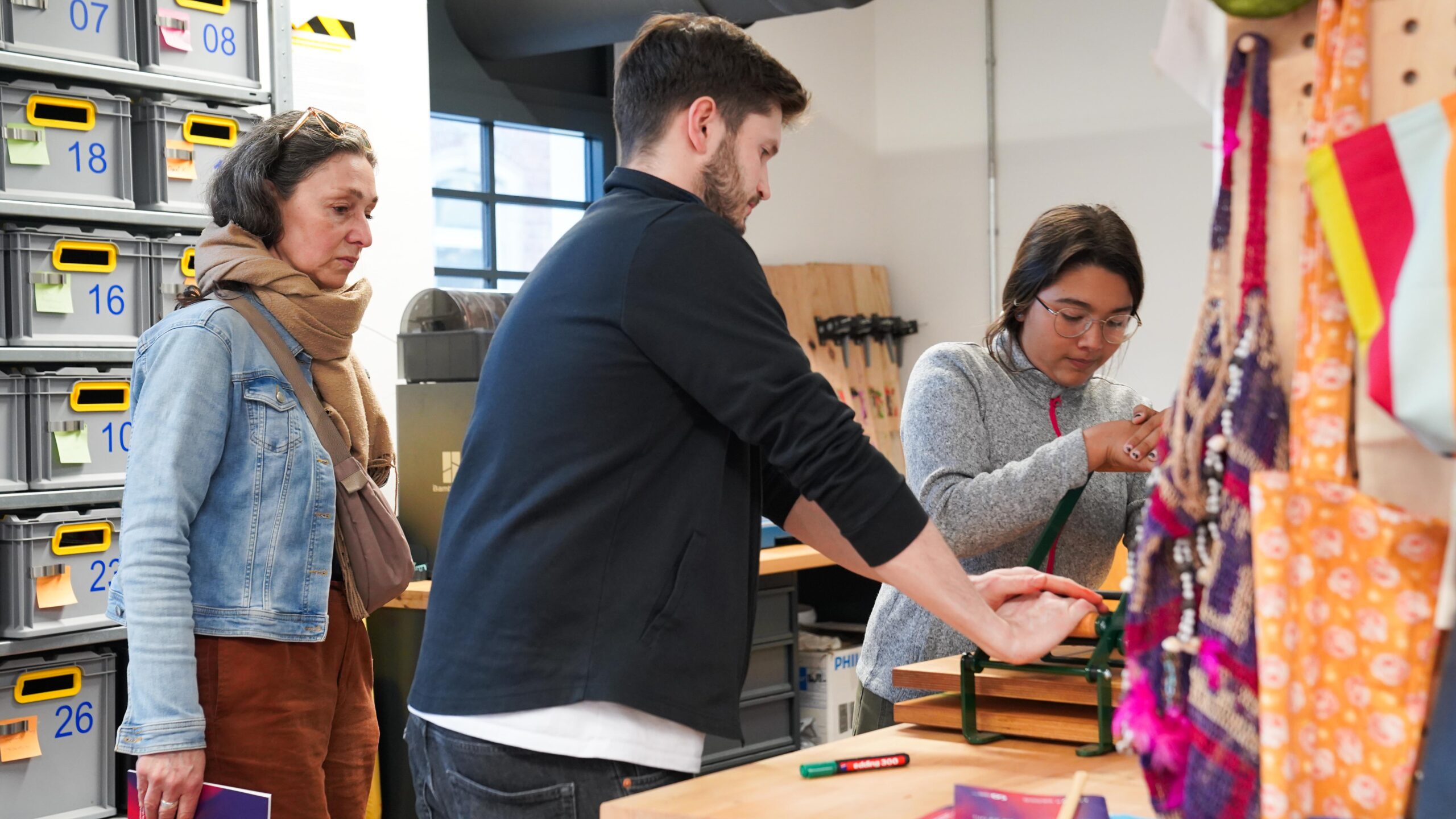 Drei Personen arbeiten an einem Tisch in einer Werkstatt während der Langen Nacht der Wissenschaften. Eine Frau beobachtet, wie ein Mann und eine andere Frau ein Werkzeug benutzen, während im Hintergrund Regale mit nummerierten Behältern und bunten Tüten zu sehen sind. at Hochschule Coburg