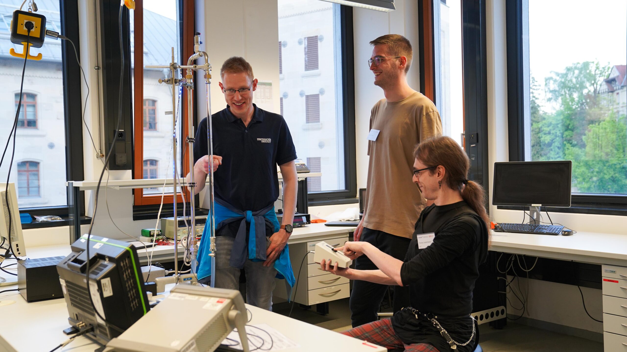 Drei Personen in einem hellen Labor mit wissenschaftlichen Geräten lächeln und interagieren während der Langen Nacht der Wissenschaften. Eine Person sitzt mit einem Gerät in der Hand, während die anderen in der Nähe stehen und neugierig auf das Experiment reagieren. at Hochschule Coburg