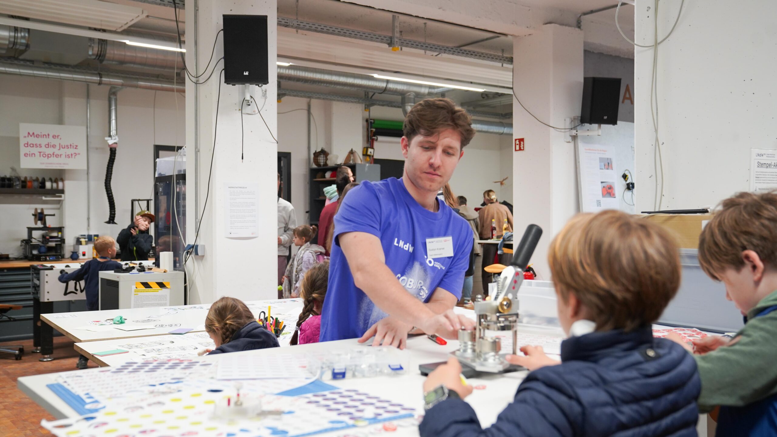 Bei der Langen Nacht der Wissenschaften hilft ein Mann in einem blauen Hemd Kindern bei einer Bastelarbeit an einem geschäftigen Werkstatttisch in einem hellen, offenen Raum. Im Hintergrund sind andere Kinder und Erwachsene zu sehen, die erste Resonanz erzeugen. at Hochschule Coburg