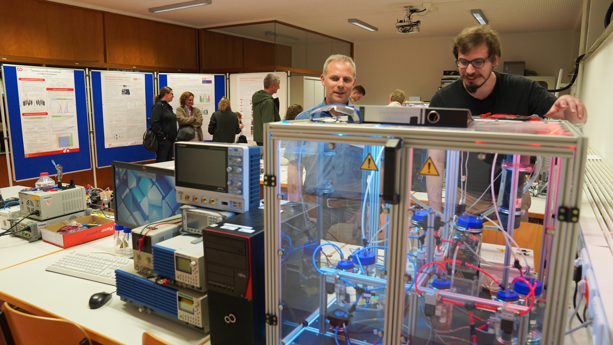 Zwei Männer untersuchen eine große Maschine mit Drähten und Schläuchen in einem Klassenzimmer während der Langen Nacht der Wissenschaften. Wissenschaftliche Geräte und Monitore füllen den Tisch, während die Besucher im Hintergrund über Plakate diskutieren und eine Atmosphäre voller Resonanz schaffen. at Hochschule Coburg