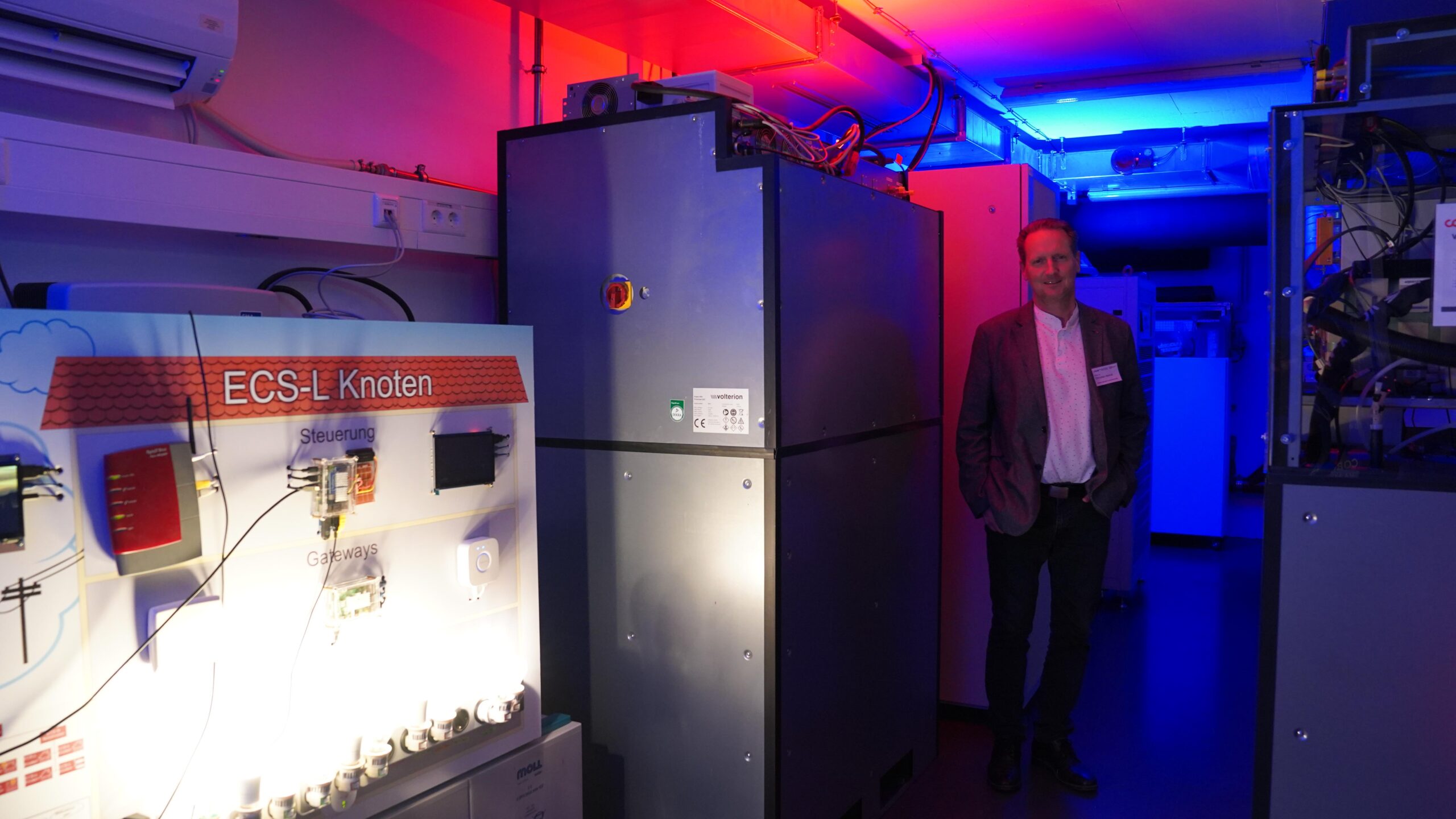 Ein Mann im Anzug steht in einem Raum mit industriellen Geräten und bunter blauer und rosa Beleuchtung neben einem Display mit der Aufschrift "ECS-L Knoten" bei der Langen Nacht der Wissenschaften, wo Wissenschaften und Resonanz im Mittelpunkt stehen. at Hochschule Coburg