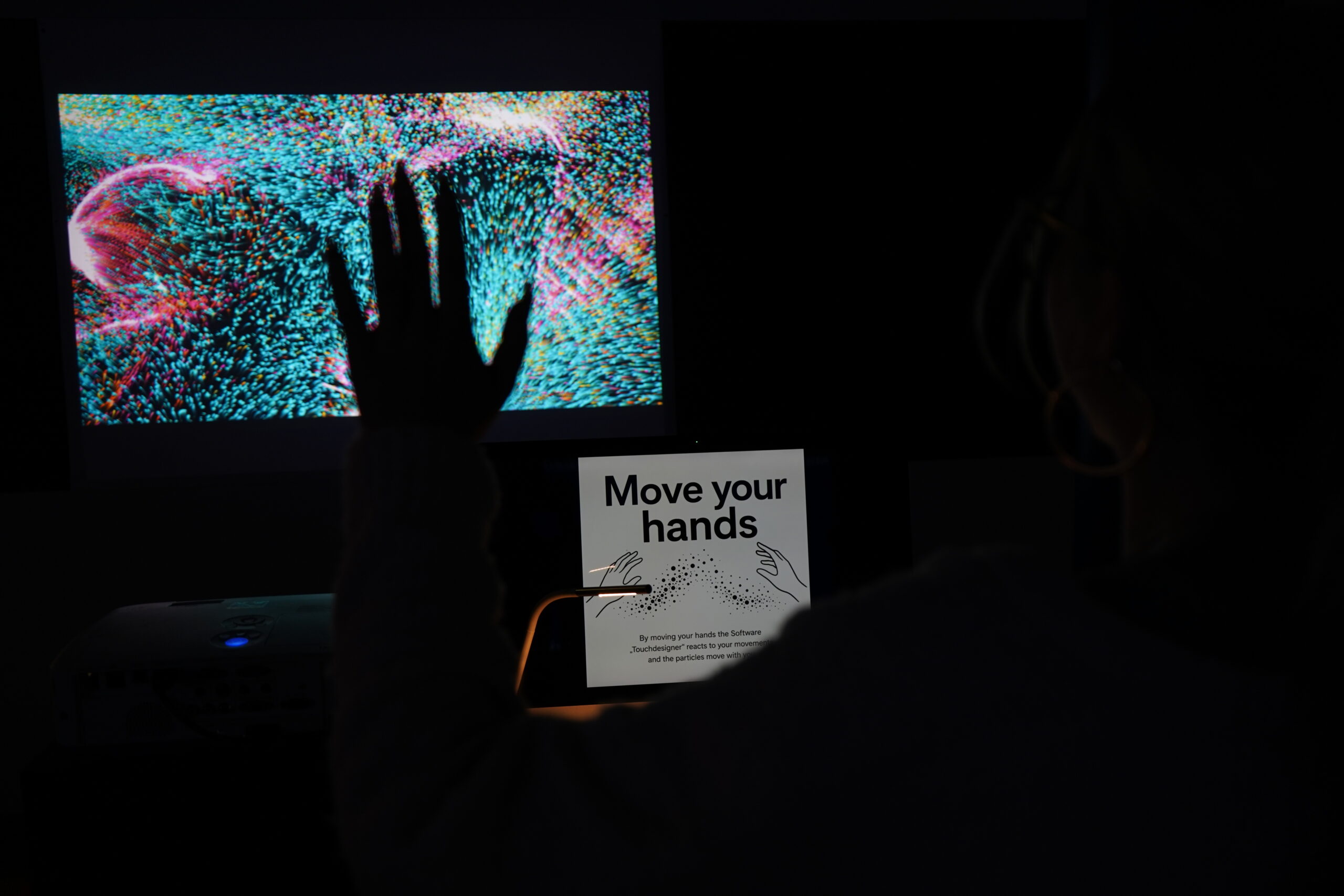 Eine Person hebt eine Hand vor einem Bildschirm mit bunten, wirbelnden Mustern in der Langen Nacht der Wissenschaften, auf dem ein Schild mit der Aufschrift "Move your hands" (Bewegen Sie Ihre Hände) und Anweisungen zur Interaktion mit dem Resonanz-Display zu lesen ist. at Hochschule Coburg