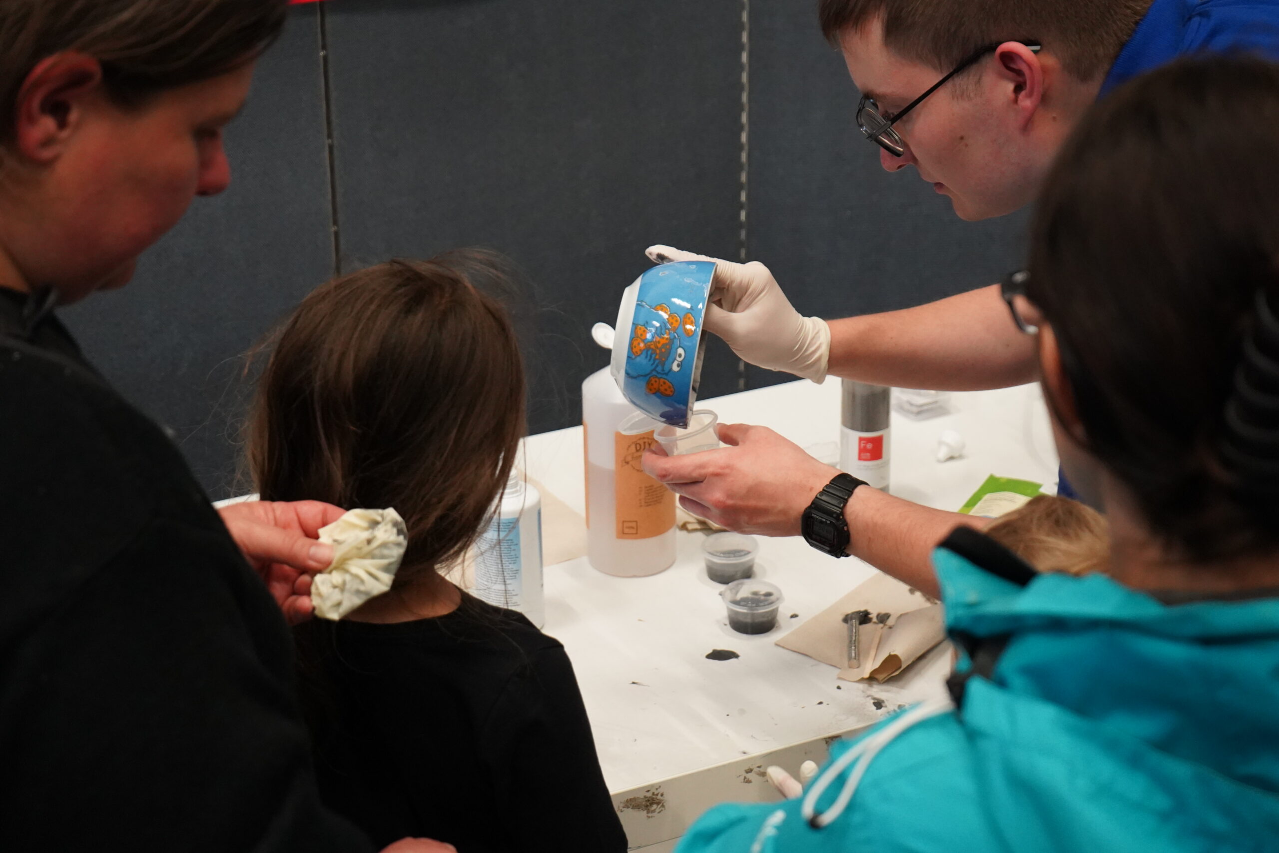 Ein Mann mit Handschuhen gießt eine Flüssigkeit aus einem Messbecher in eine Flasche, während zwei Kinder und ein Erwachsener an einem weißen Tisch, der mit Materialien aus dem Bereich Wissenschaften bedeckt ist, aufmerksam zusehen und so den neugierigen Geist der Langen Nacht der Wissenschaften einfangen. at Hochschule Coburg