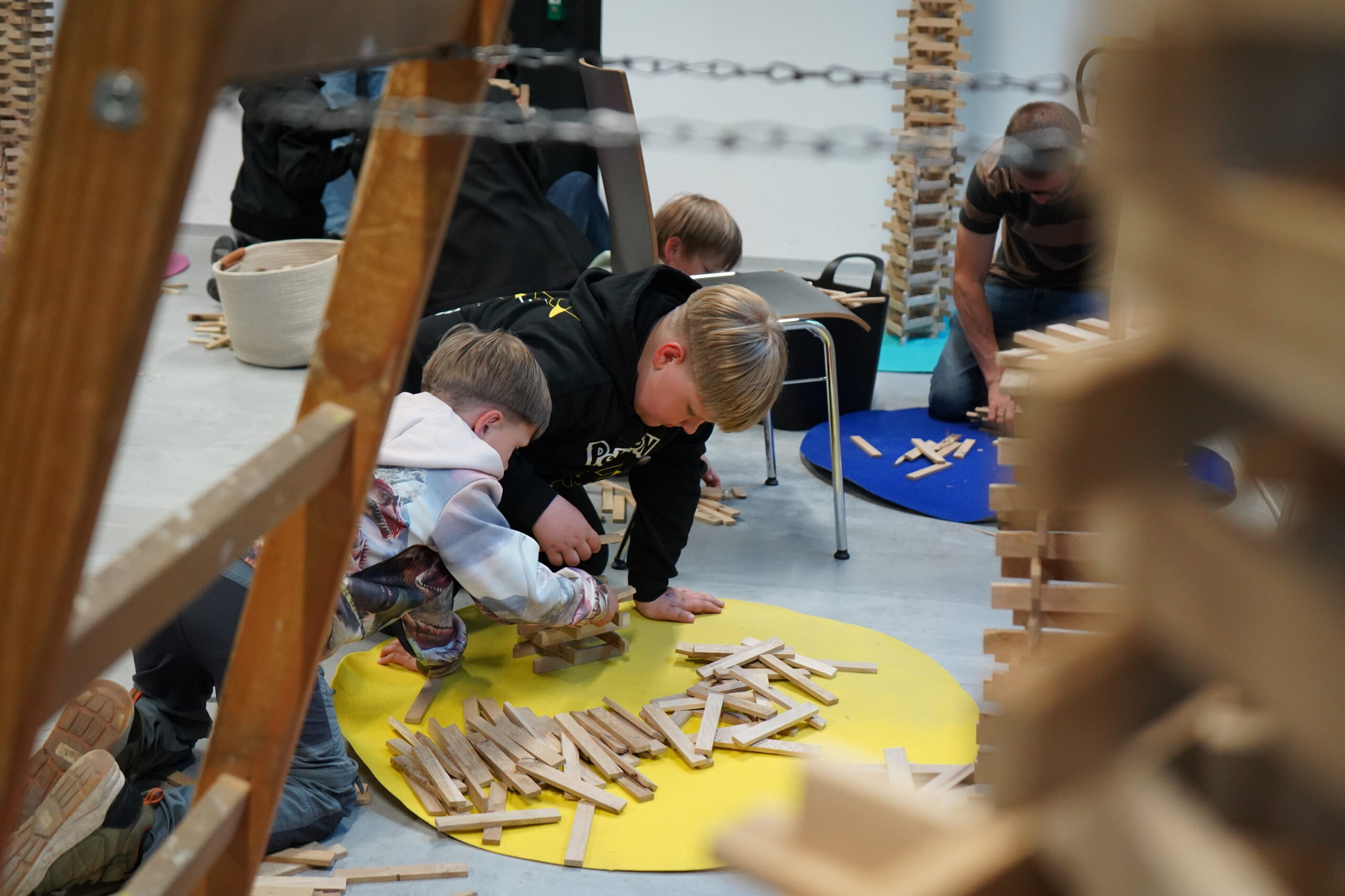 Zwei Jungen knien auf einer gelben Matte und bauen mit Holzklötzen Strukturen, die Resonanz und Neugierde wecken. Ein Erwachsener und ein weiteres Kind im Hintergrund spielen ebenfalls und schaffen eine lebendige, von der Wissenschaft inspirierte Atmosphäre, die an die Lange Nacht der Wissenschaften erinnert. at Hochschule Coburg