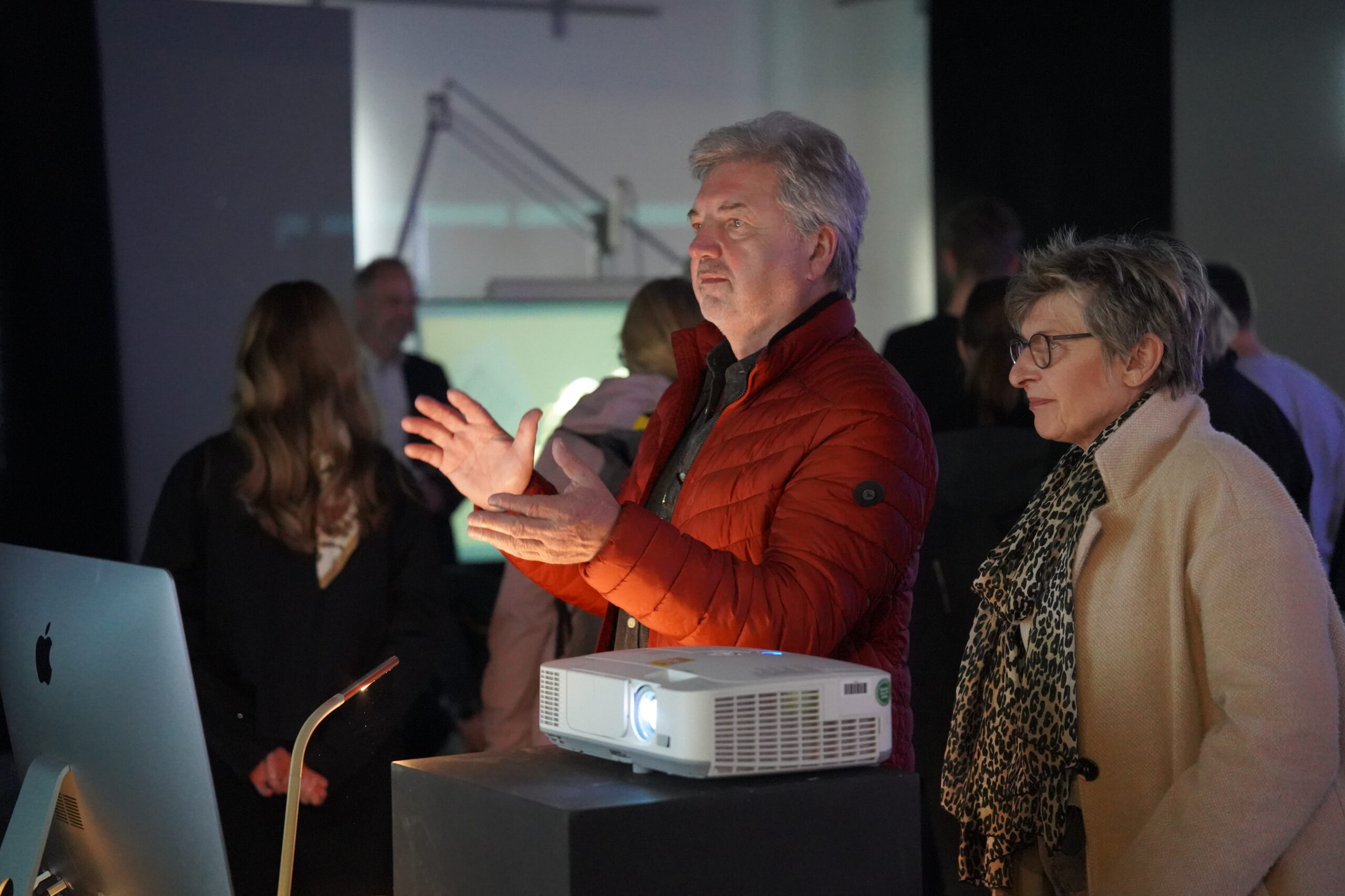 Ein Mann in einer roten Jacke gestikuliert, während er neben einer Frau mit Brille und braunem Mantel steht. Vor ihnen befinden sich ein Projektor und ein Computer bei der Langen Nacht der Wissenschaften, während mehrere Personen im Hintergrund zusehen. at Hochschule Coburg