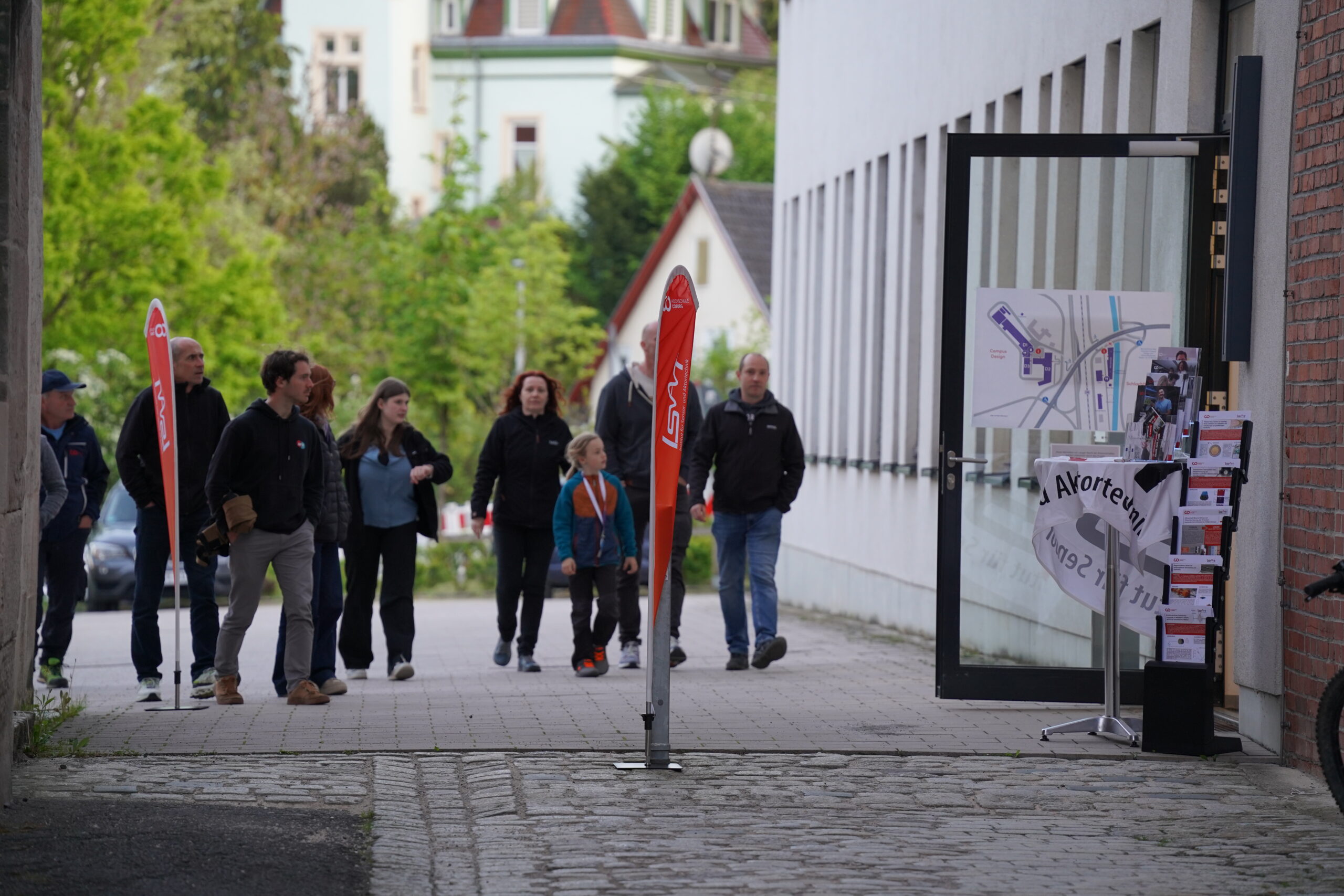 Eine Gruppe von Menschen geht einen gepflasterten Weg entlang zu einer offenen Glastür, an der Informationsplakate und eine große Karte für die Lange Nacht der Wissenschaften angebracht sind. Grüne Bäume und Gebäude bilden den Hintergrund. at Hochschule Coburg