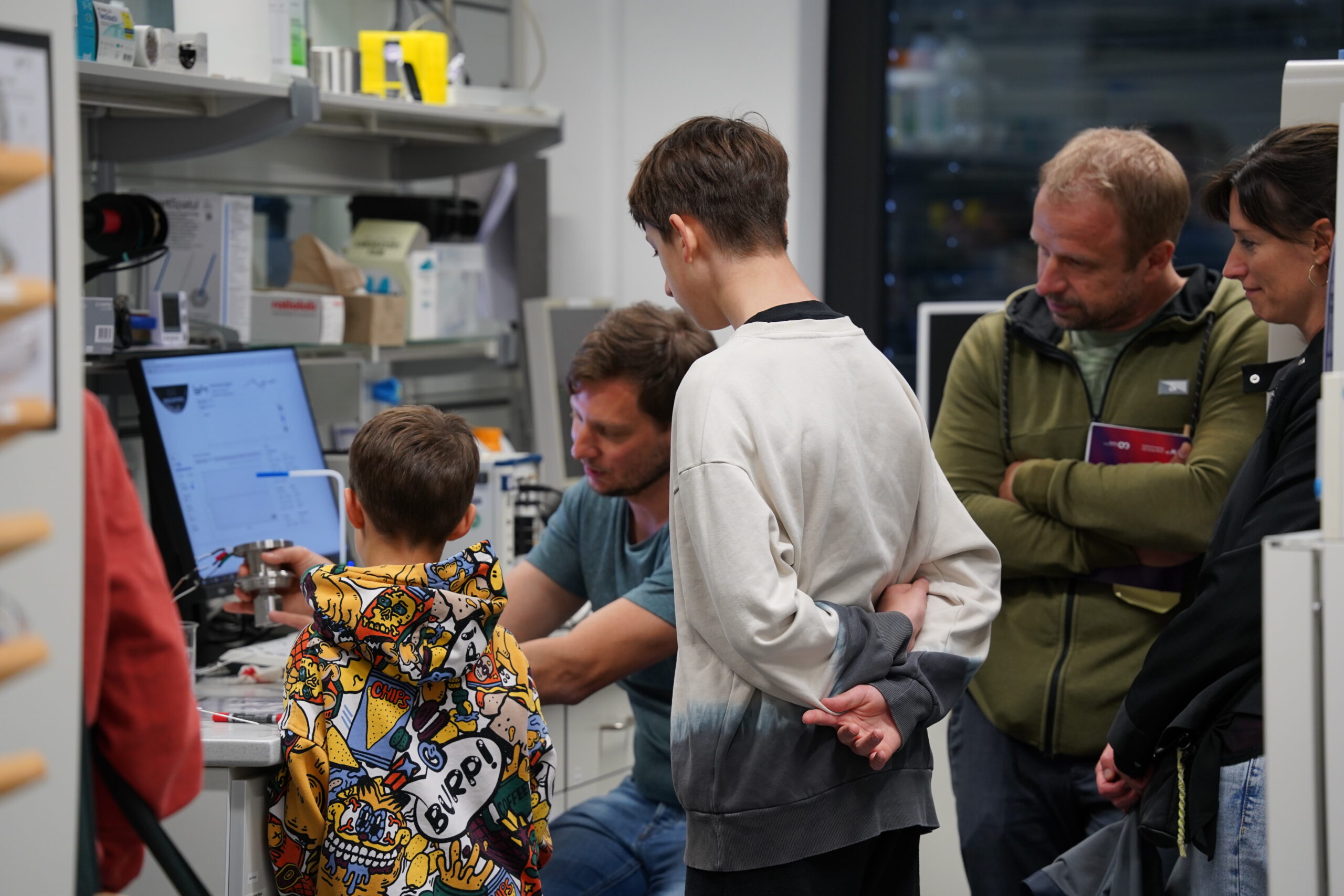 Während der Langen Nacht der Wissenschaften versammelt sich eine Gruppe von Kindern und Erwachsenen um einen Wissenschaftler in einem Labor voller Geräte und lässt sich von einer Computervorführung fesseln, die bei den neugierigen Teilnehmern Resonanz auslöst. at Hochschule Coburg