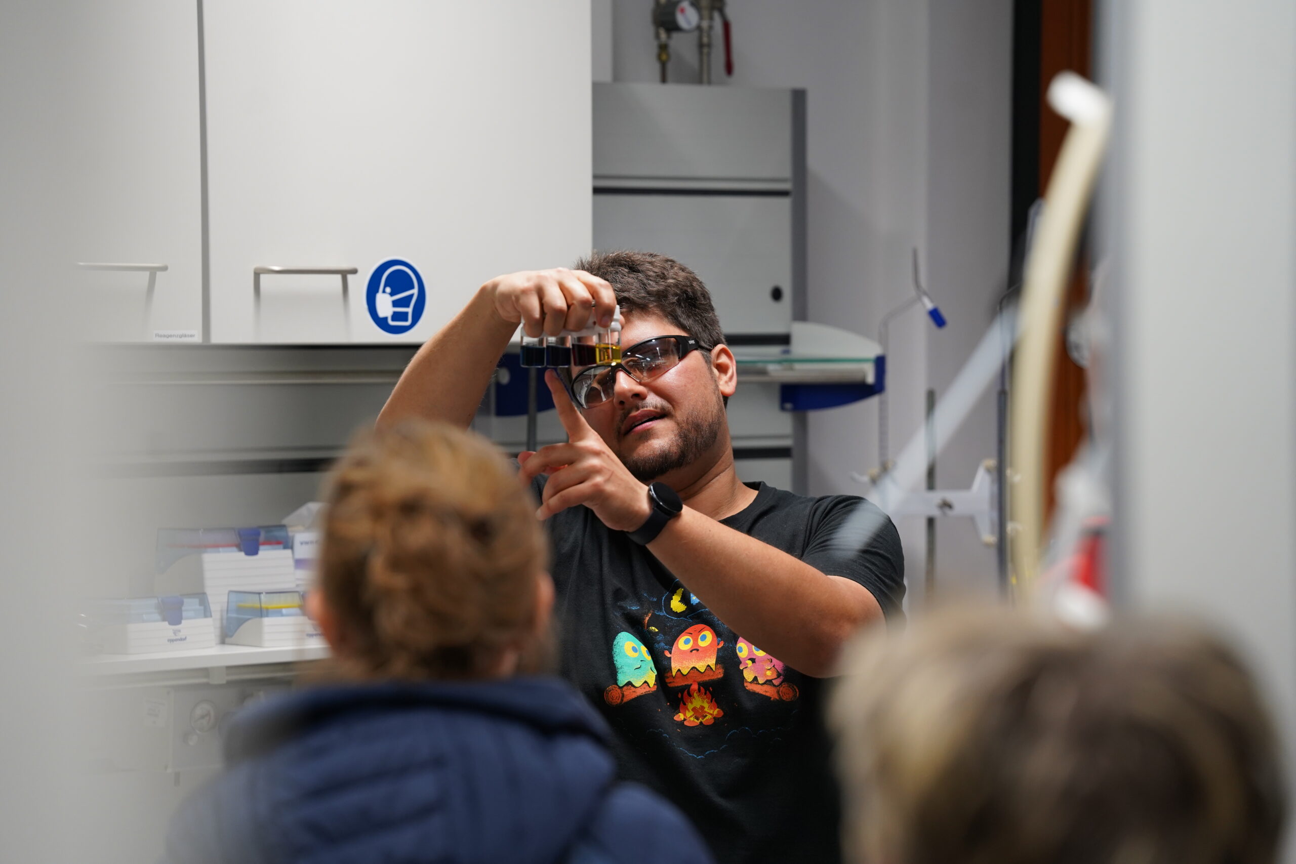 Auf einer Wissenschaftsveranstaltung während der Langen Nacht der Wissenschaften hält ein Mann mit Sonnenbrille und einem bunten Hemd mit Cartoon-Monstern in einem Labor ein Fläschchen hoch und demonstriert zwei Personen, die mit dem Rücken zur Kamera stehen, etwas. at Hochschule Coburg