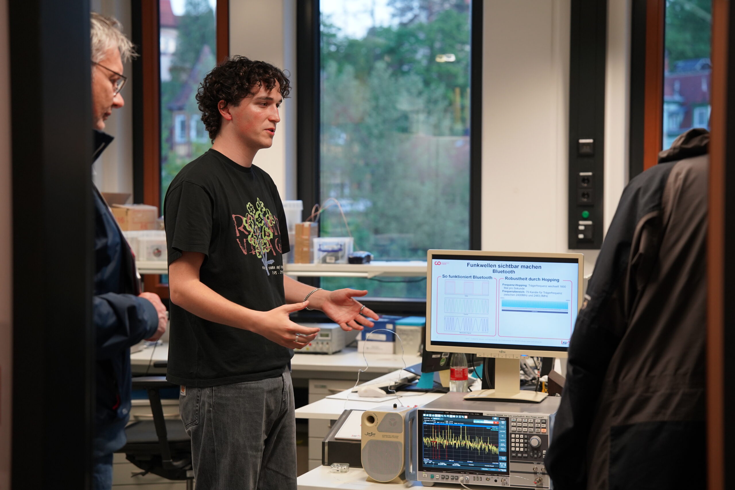 Ein junger Mann steht vor einem Computermonitor mit Diagrammen und erklärt zwei anderen in einem modernen Labor etwas über Wissenschaft. Auf dem Schreibtisch sind elektronische Geräte und Kabel zu sehen, die den Geist der Langen Nacht der Wissenschaften einfangen. at Hochschule Coburg