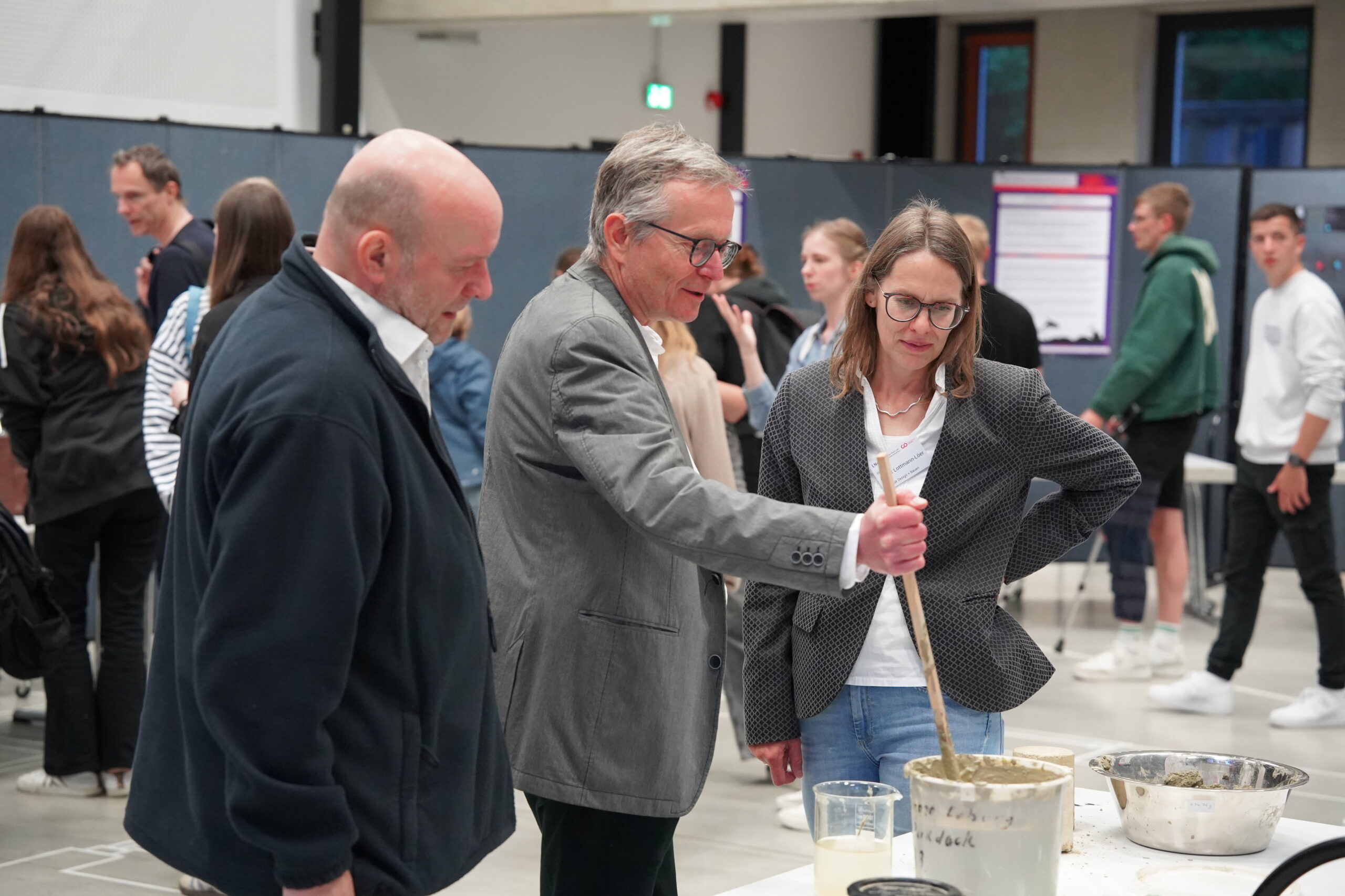 Drei Erwachsene stehen um einen Tisch mit Schüsseln und einem großen Behälter, eine Frau rührt eine Mischung um. Im Hintergrund sind weitere Personen bei der Langen Nacht der Wissenschaften zu sehen, was auf eine öffentliche wissenschaftliche Veranstaltung in einer geräumigen Halle hindeutet. at Hochschule Coburg
