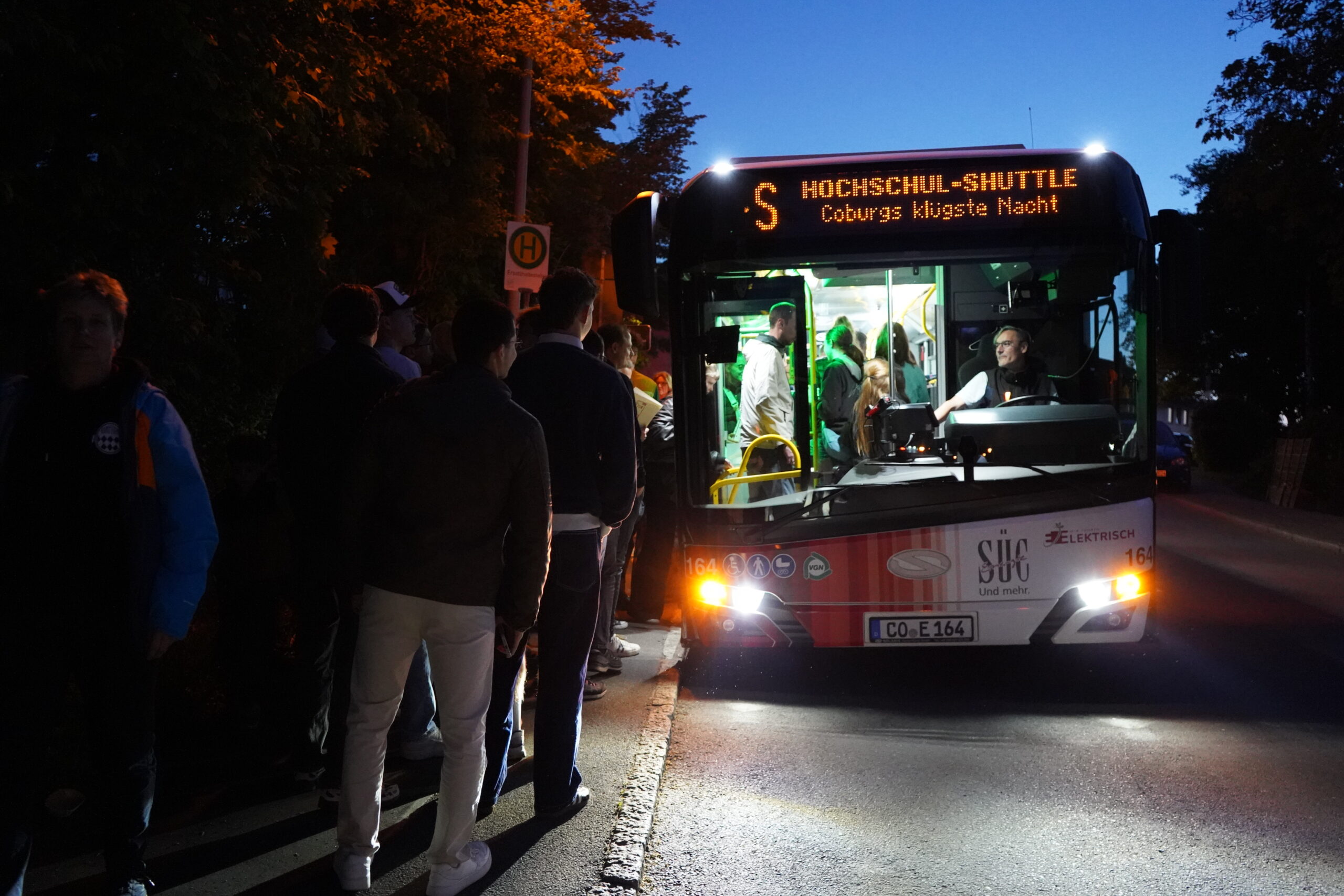 Eine Gruppe von Menschen besteigt nachts einen hell erleuchteten Hochschul-Shuttle-Bus für die Lange Nacht der Wissenschaften. Auf dem Busschild steht "Coburgs klügste Nacht", beleuchtet von Straßenlampen und Scheinwerfern, die eine Atmosphäre wissenschaftlicher Resonanz schaffen. at Hochschule Coburg