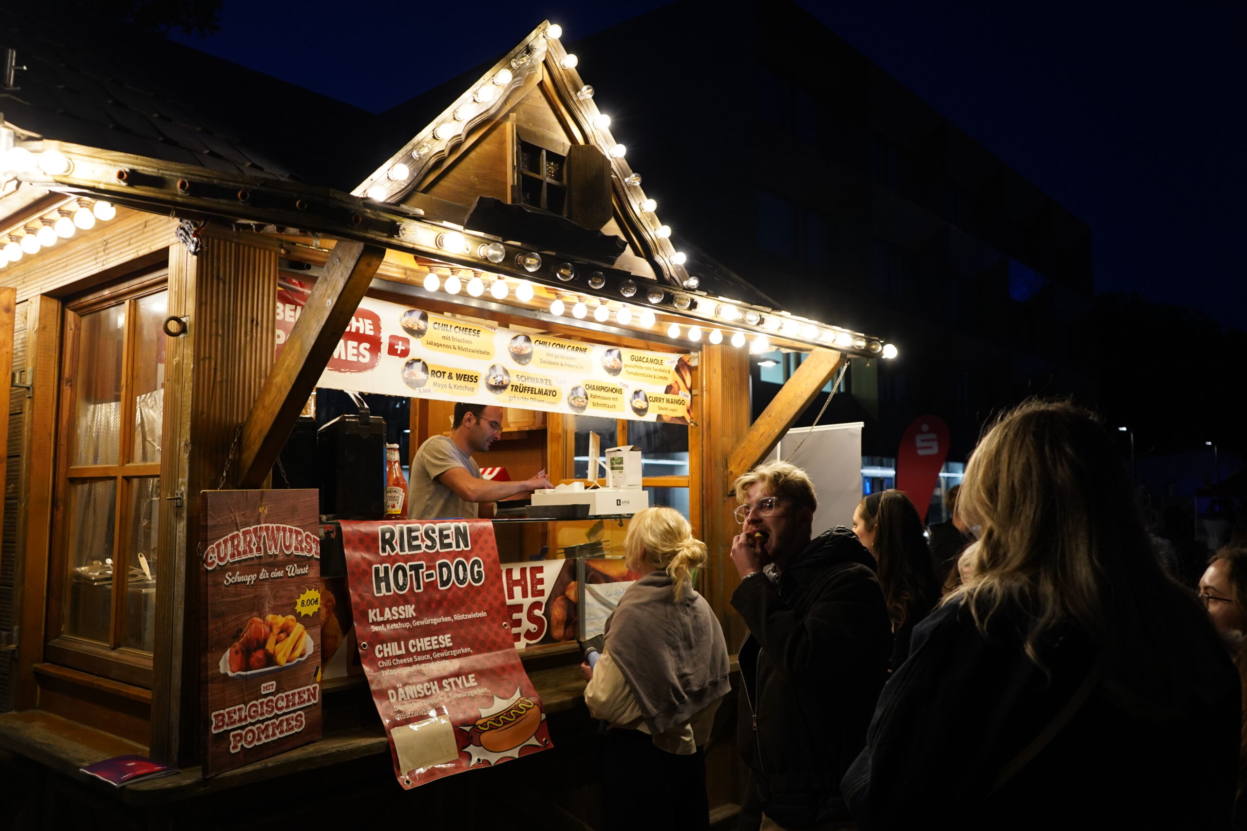 Eine Gruppe von Menschen steht während der Langen Nacht der Wissenschaften vor einem hell erleuchteten hölzernen Essensstand an. Der Stand wirbt mit bunten Schildern für Hot Dogs und Pommes Frites, während ein Verkäufer im Inneren das Essen zubereitet. at Hochschule Coburg