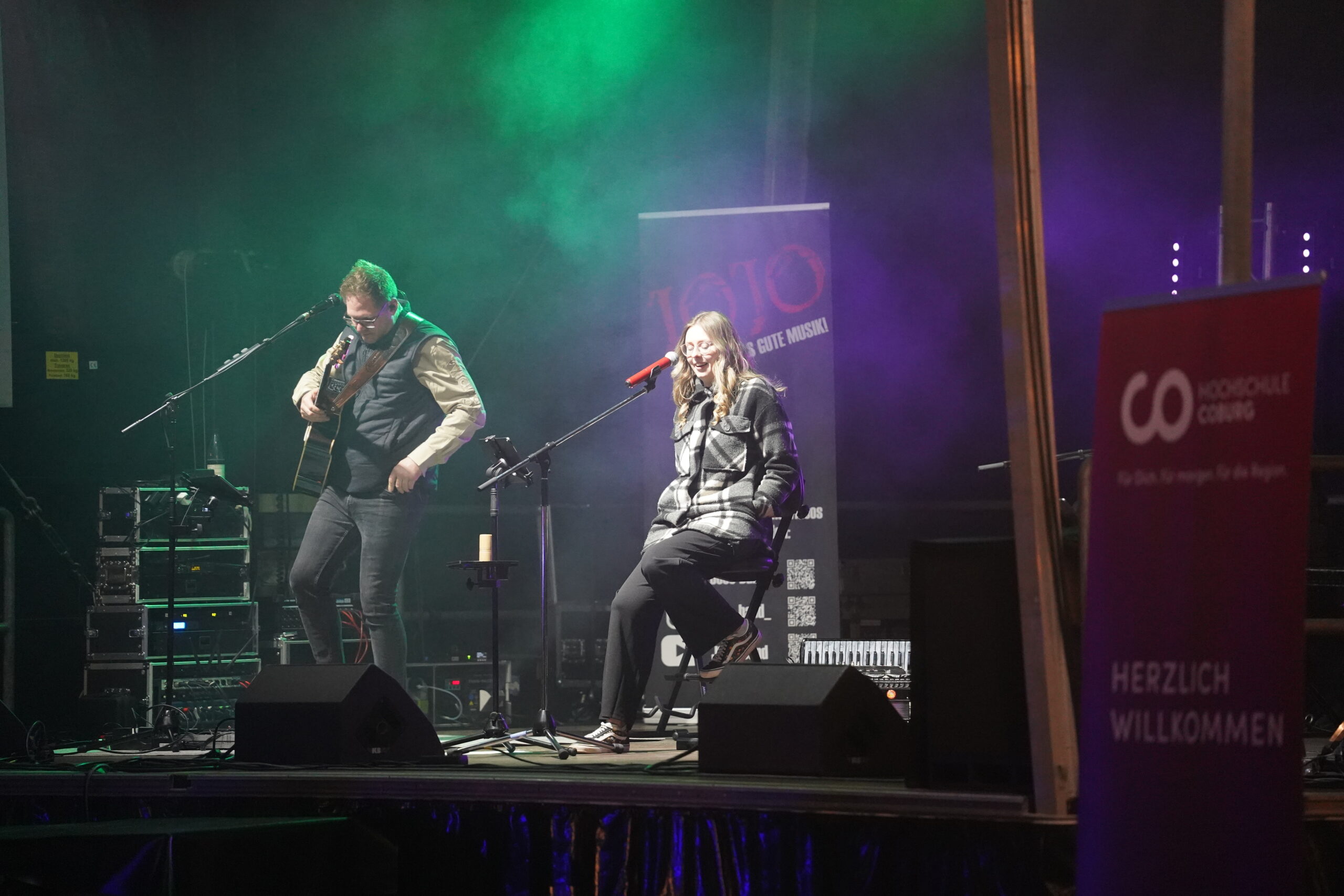 Zwei Musiker treten auf einer schwach beleuchteten Bühne bei der Langen Nacht der Wissenschaften auf, einer spielt Gitarre, der andere singt in ein Mikrofon. Farbige Lichter, Nebeleffekte und ein "Herzlich Willkommen"-Schild sorgen für eine stimmungsvolle Atmosphäre. at Hochschule Coburg