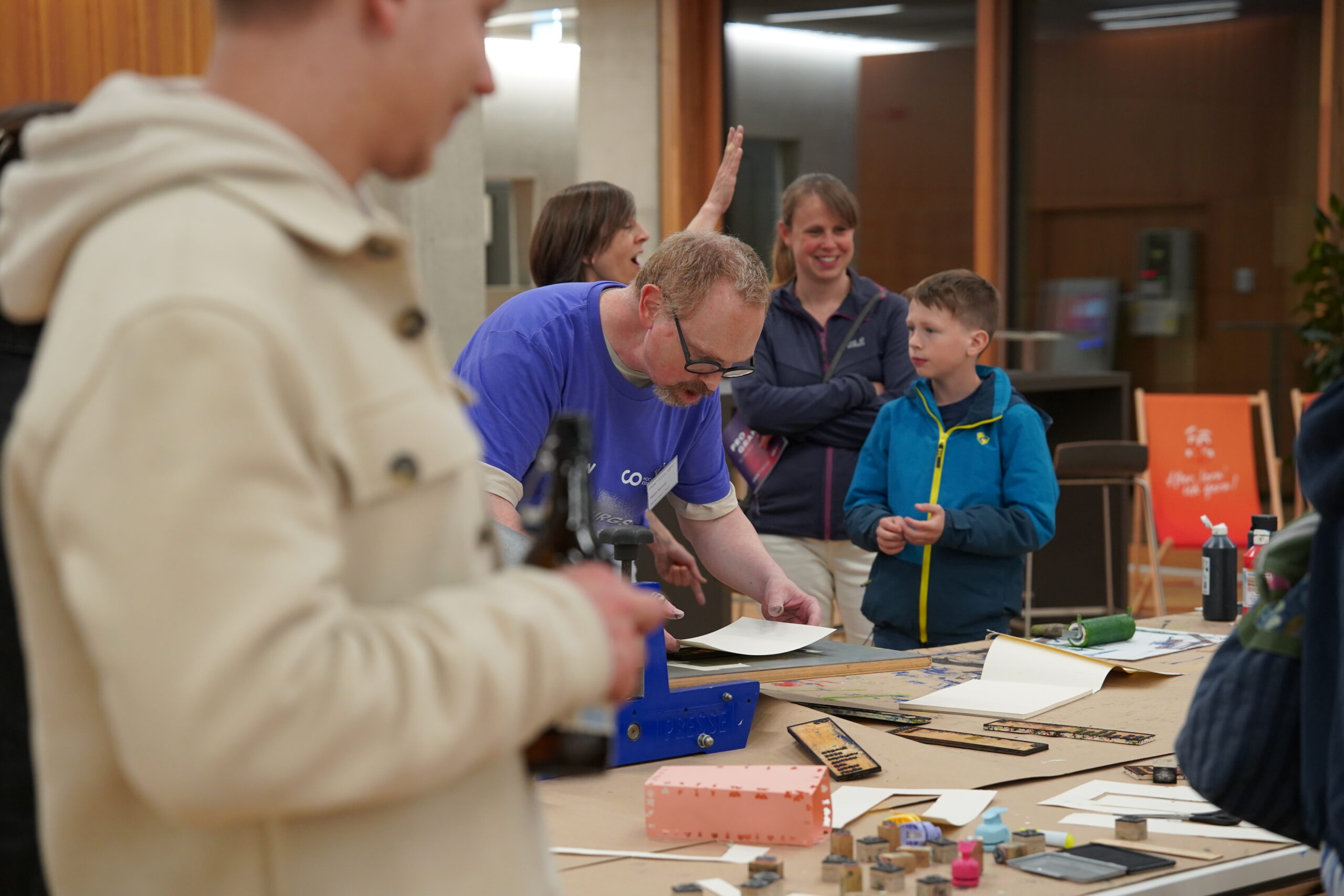 Ein Mann in einem blauen Hemd führt während der Langen Nacht der Wissenschaften an einem Tisch mit Kunstzubehör eine Bastelarbeit vor, während mehrere Erwachsene und Kinder in einem hell erleuchteten Innenraum zusehen und interagieren und so eine Atmosphäre voller Resonanz schaffen. at Hochschule Coburg