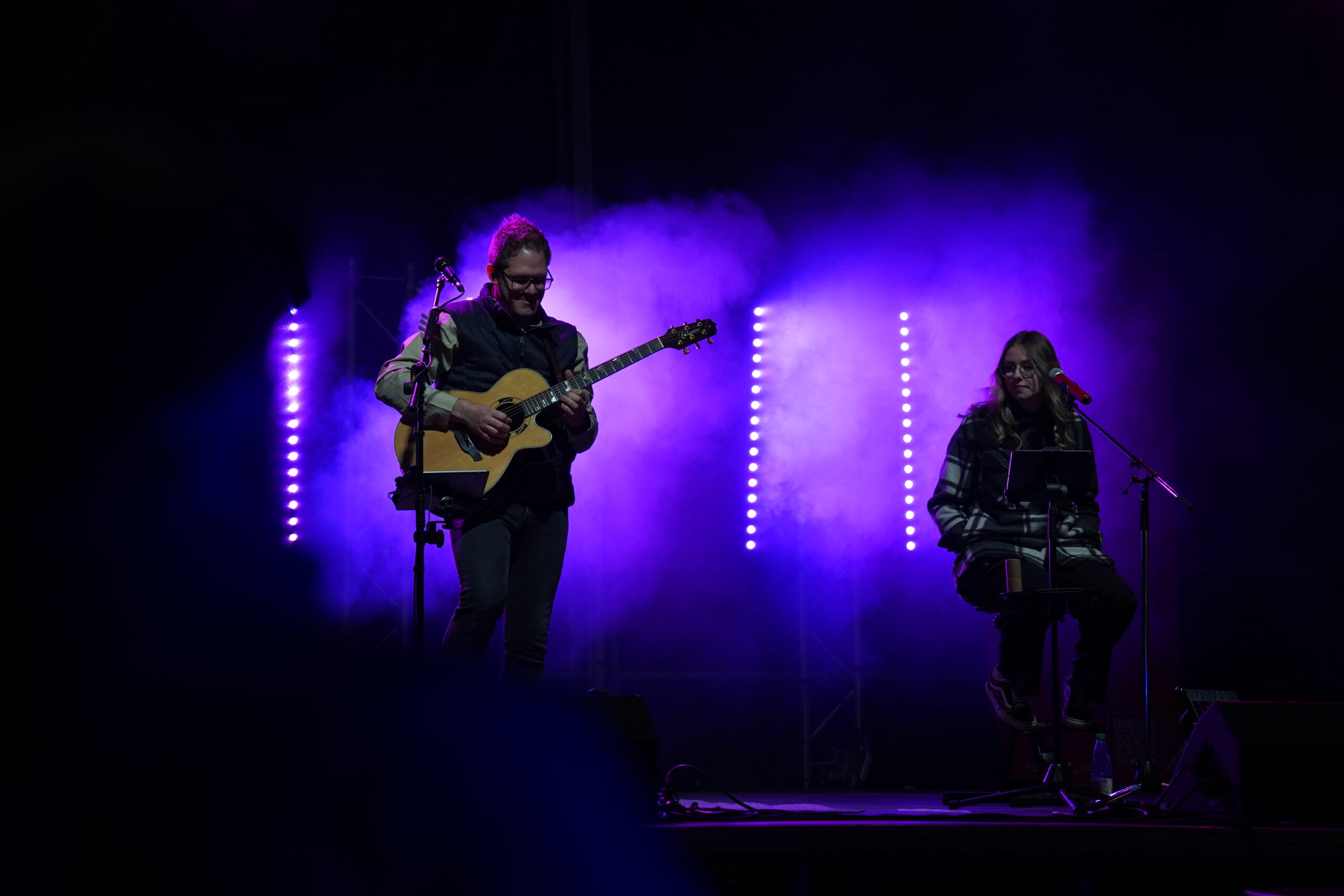 Zwei Musiker treten auf der Bühne unter violettem Licht und Nebel auf, was ein Gefühl von Resonanz erzeugt; einer spielt Gitarre und singt, während der andere mit Mikrofon und Notenständer in der Langen Nacht der Wissenschaften sitzt. at Hochschule Coburg