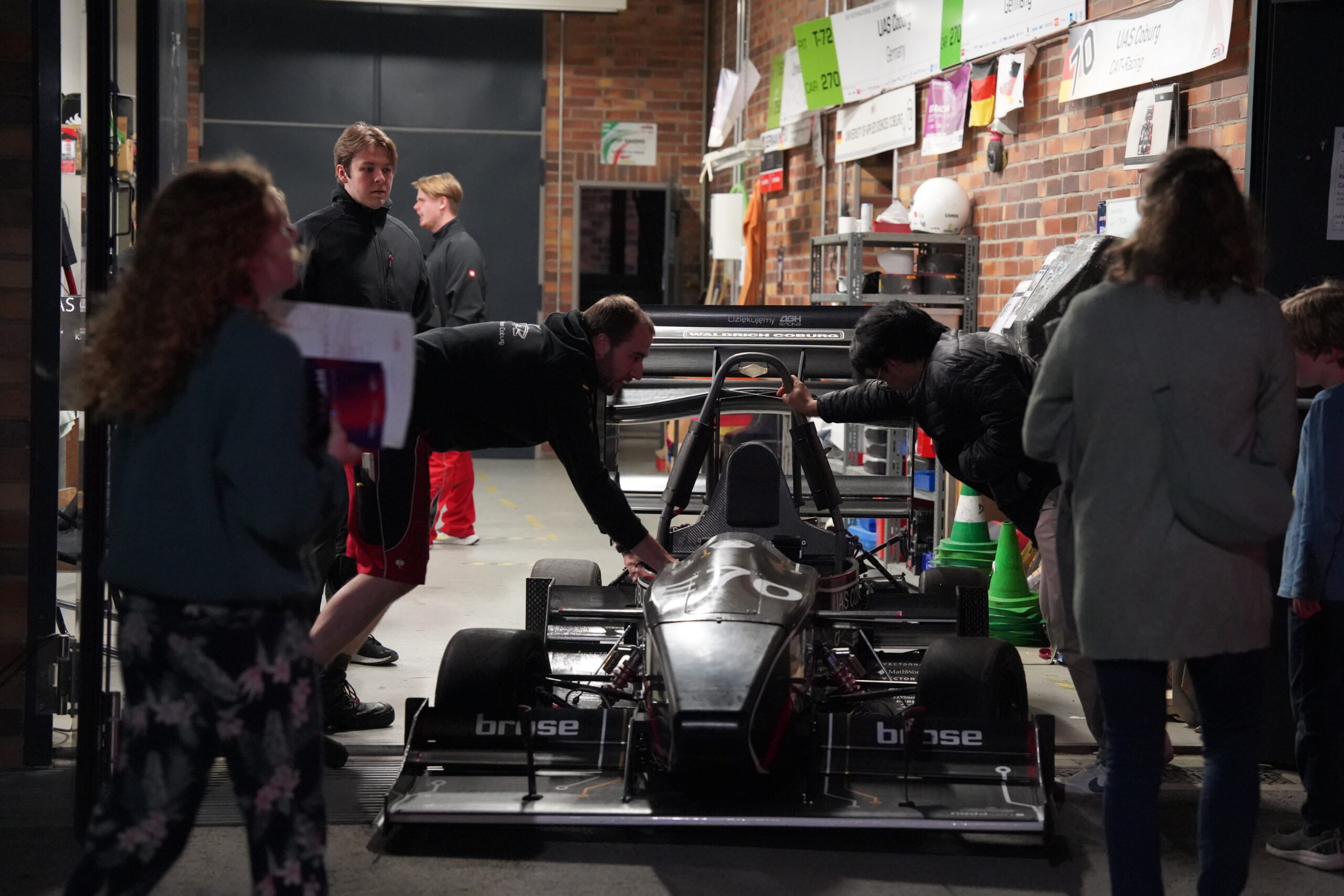 Eine Gruppe von Menschen steht in einer Garage während der Langen Nacht der Wissenschaften um einen schwarzen Rennwagen mit offenem Lenkrad herum und inspiziert ihn, umgeben von Werkzeugen, Bannern und Geräten. Die lebhafte Atmosphäre spiegelt die Resonanz von Neugierde und Innovation wider. at Hochschule Coburg
