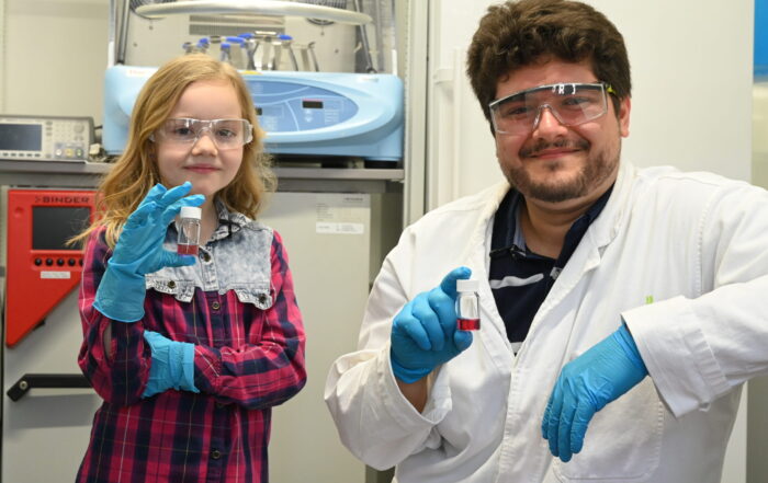 Ein junges Mädchen und ein erwachsener Mann, beide mit Schutzbrille und blauen Handschuhen, halten in einer Laborumgebung kleine Fläschchen und lächeln in die Kamera. Im Hintergrund sind Laborgeräte und wissenschaftliche Instrumente zu sehen. at Hochschule Coburg