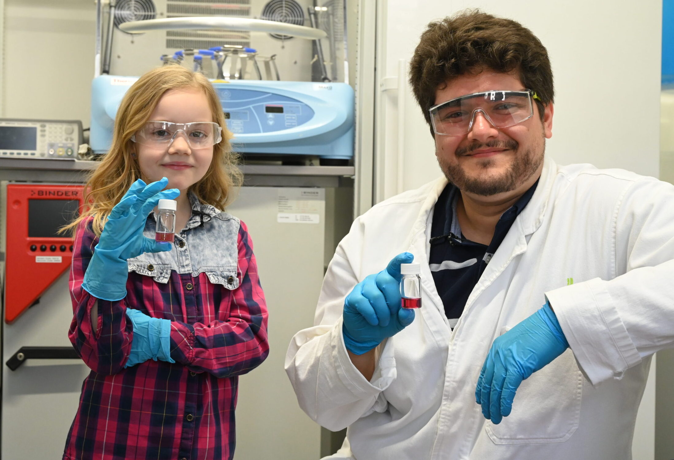 Ein junges Mädchen und ein erwachsener Mann, beide mit Schutzbrille und blauen Handschuhen, halten in einer Laborumgebung kleine Fläschchen und lächeln in die Kamera. Im Hintergrund sind Laborgeräte und wissenschaftliche Instrumente zu sehen. at Hochschule Coburg