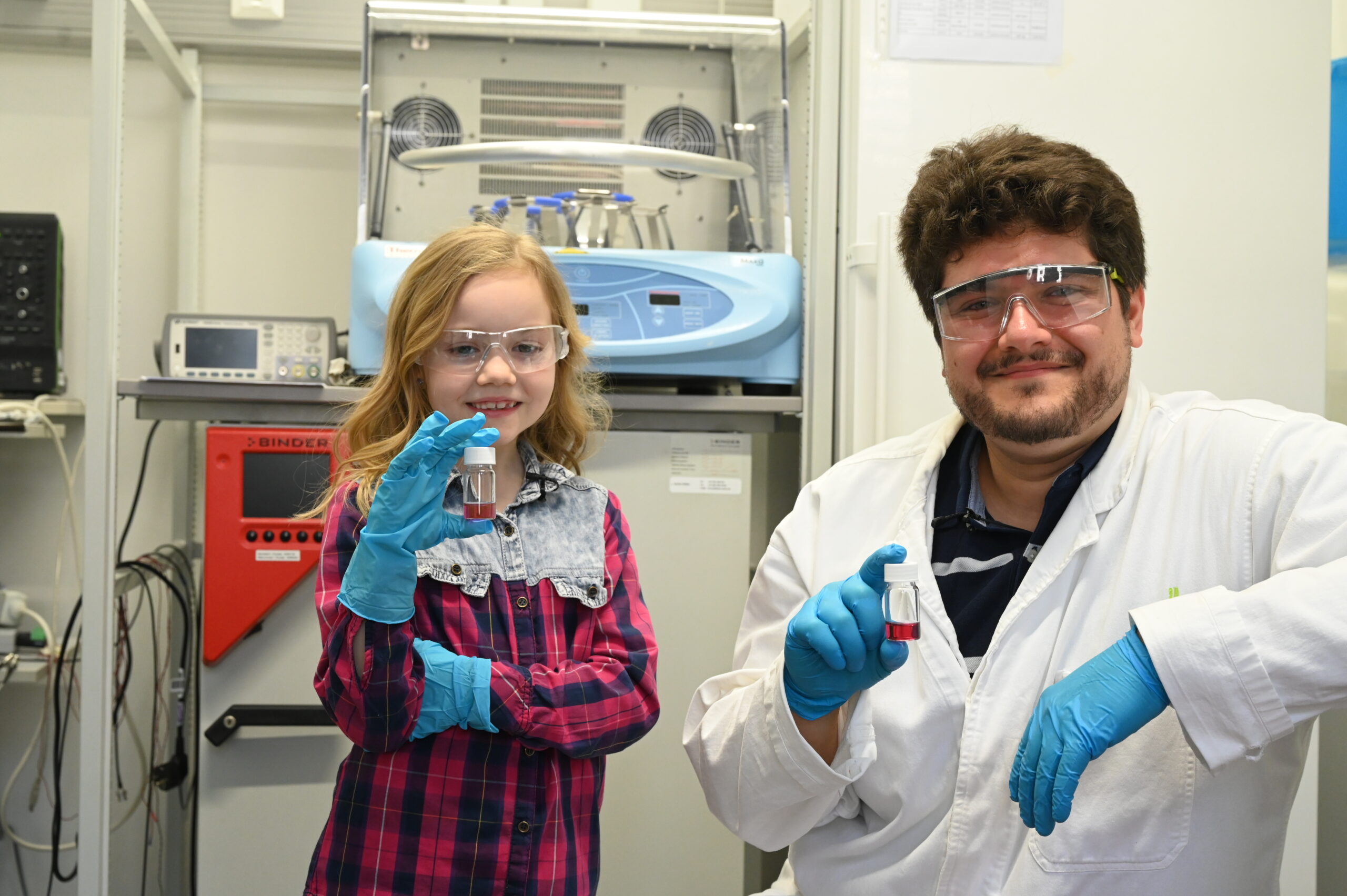Ein junges Mädchen und ein Mann in Laborkittel und Handschuhen, beide mit Schutzbrille, halten kleine Fläschchen in einer Laborumgebung mit wissenschaftlichen Geräten im Hintergrund. at Hochschule Coburg