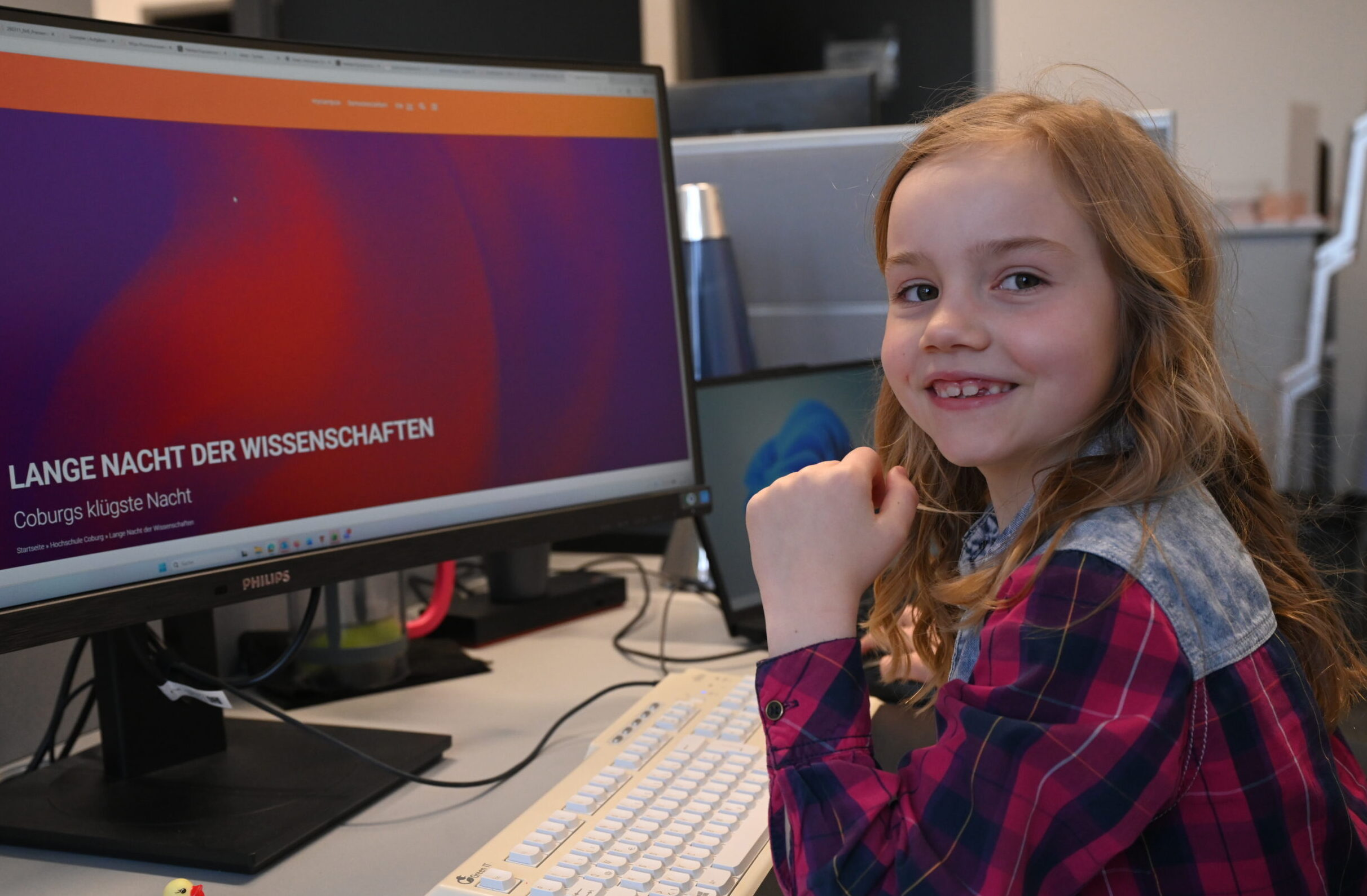 Ein lächelndes junges Mädchen mit langen blonden Haaren sitzt an einem Schreibtisch und benutzt einen Computer. Auf dem Monitor erscheint eine Website mit dem deutschen Text "Lange Nacht der Wissenschaften". at Hochschule Coburg