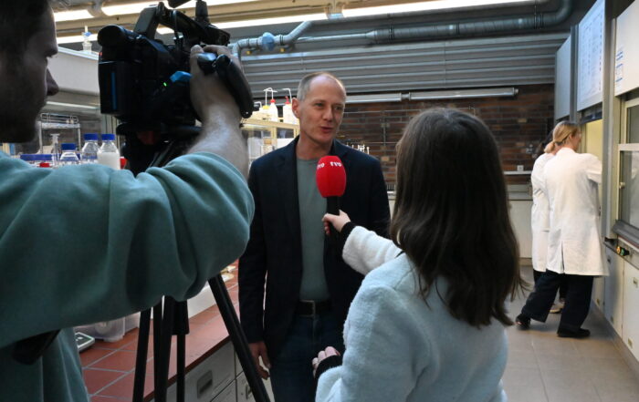 Ein Mann in einem Blazer wird mit einem roten TVO-Mikrofon interviewt und von einem Kameramann in einem Labor während der LNdW gefilmt, während im Hintergrund eine Person in einem weißen Laborkittel arbeitet. at Hochschule Coburg