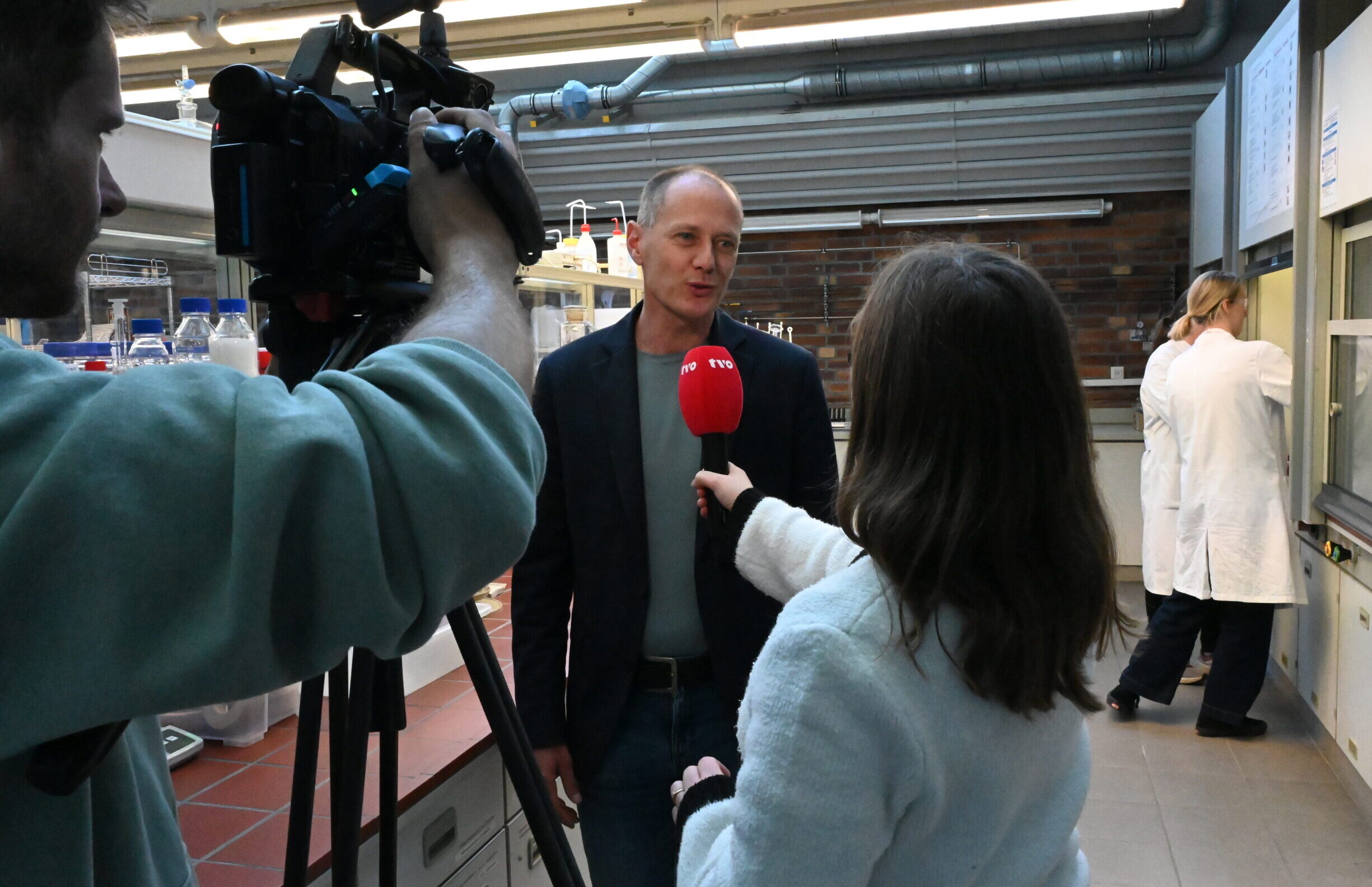Ein Mann in einem Blazer wird mit einem roten TVO-Mikrofon interviewt und von einem Kameramann in einem Labor während der LNdW gefilmt, während im Hintergrund eine Person in einem weißen Laborkittel arbeitet. at Hochschule Coburg