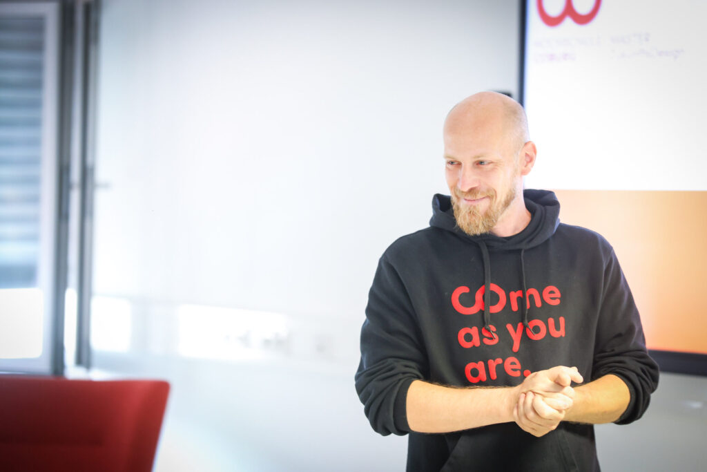 Ein lächelnder Mann mit Bart, der einen schwarzen Kapuzenpulli mit der Aufschrift "Come as you are" trägt, steht in einem hellen Raum neben einer Projektionsfläche und strahlt den Geist von Innovation und Unternehmertum aus. at Hochschule Coburg