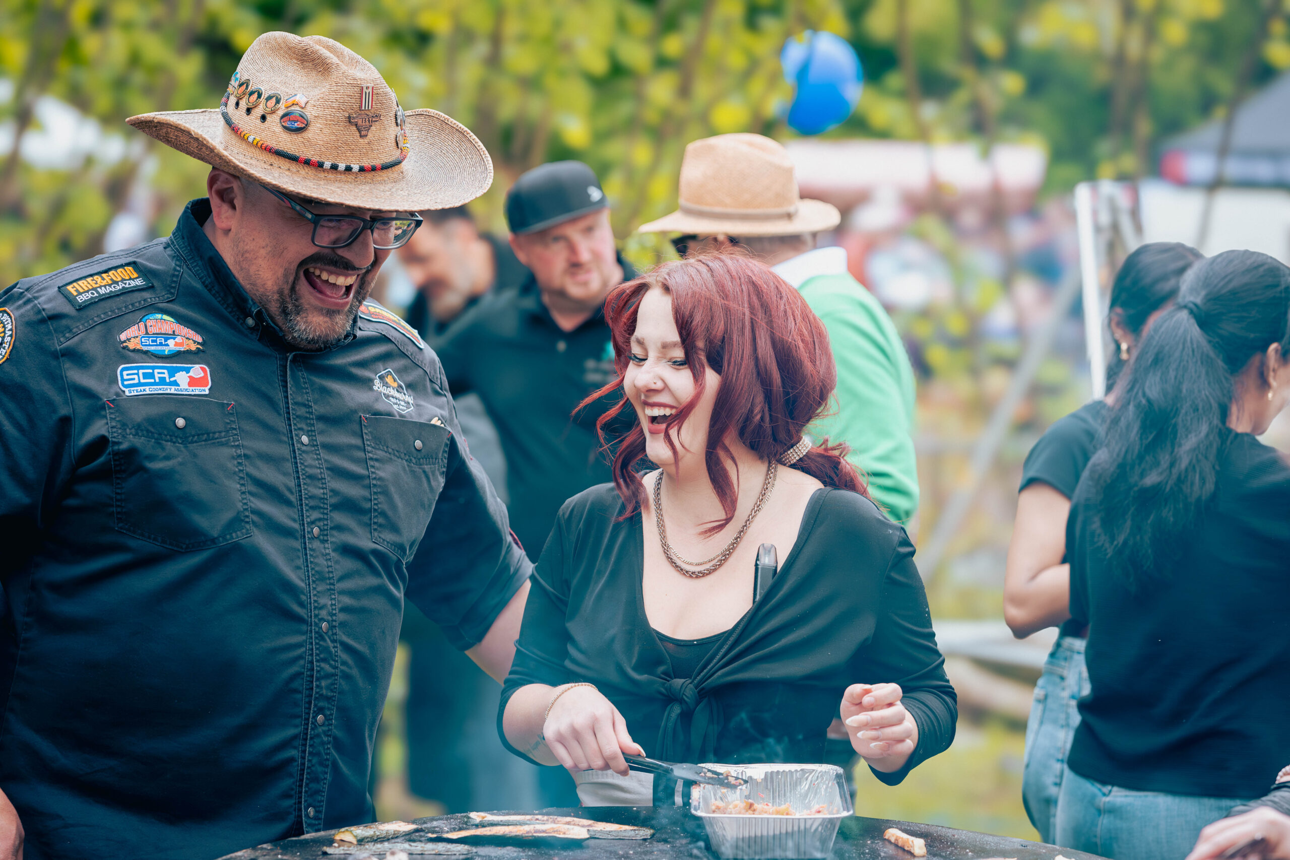 Zwei Personen lächeln und lachen, während sie auf der Hochschul-Grillmeisterschaft im Freien grillen. Der Mann trägt einen Cowboyhut und ein verziertes Hemd, während die rothaarige Frau mit einer Zange Tofugerichte grillt, während sich andere im Hintergrund unter sie mischen. at Hochschule Coburg