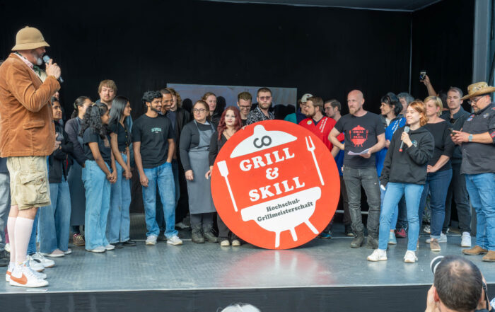 Eine Gruppe auf der Bühne steht um ein "Grill & Skill Hochschul-Grillmeisterschaft"-Schild herum, während ein Mann mit einem Mikrofon zu ihnen spricht. Lächelnde Teilnehmer schauen zu und freuen sich auf die Hochschul-Grillmeisterschaft 2026 und kreative Gerichte wie Zucchini-Rettung. at Hochschule Coburg