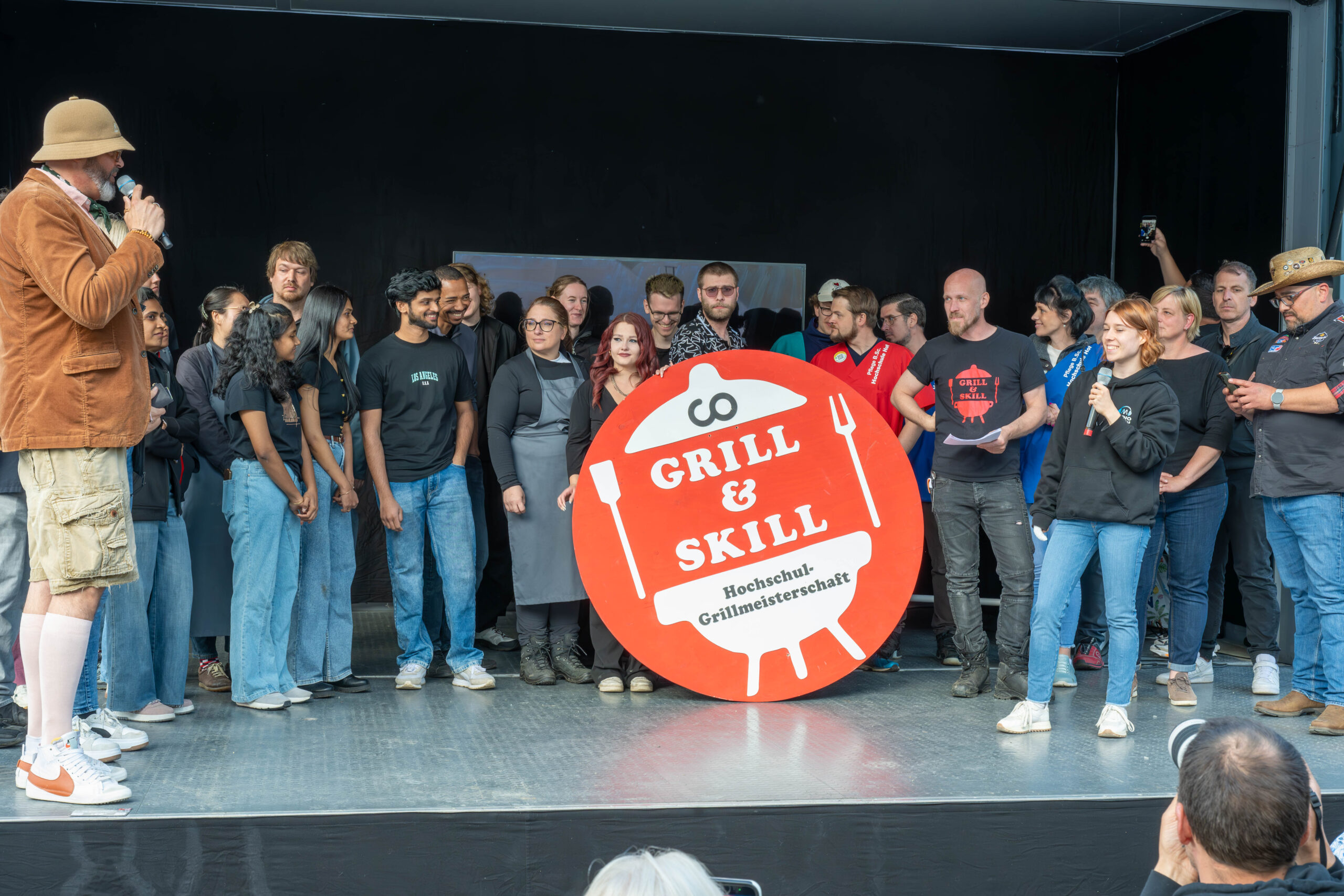 Eine Gruppe auf der Bühne steht um ein "Grill & Skill Hochschul-Grillmeisterschaft"-Schild herum, während ein Mann mit einem Mikrofon zu ihnen spricht. Lächelnde Teilnehmer schauen zu und freuen sich auf die Hochschul-Grillmeisterschaft 2026 und kreative Gerichte wie Zucchini-Rettung. at Hochschule Coburg
