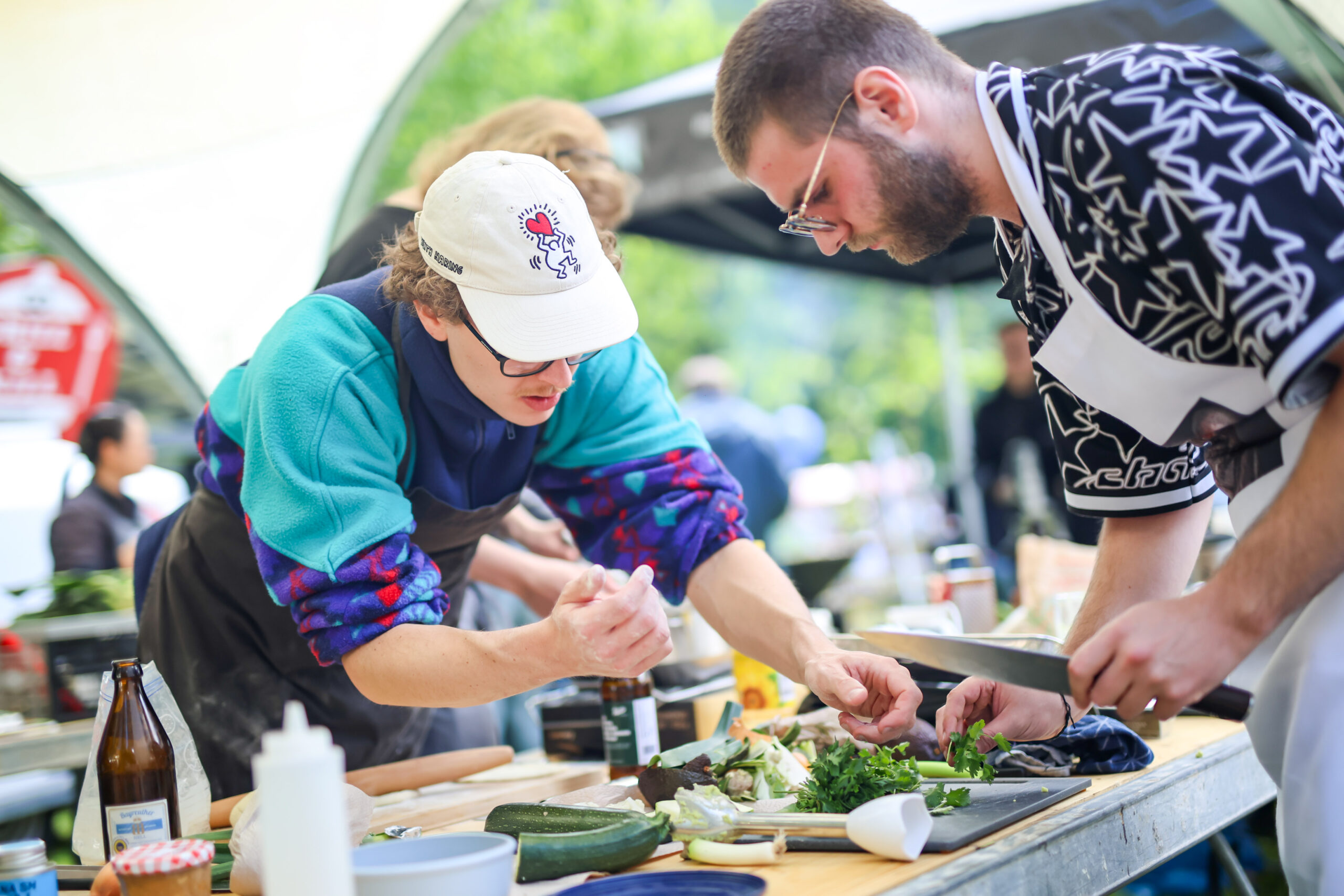 Zwei Personen in Schürzen bereiten gemeinsam im Freien das Essen zu. Konzentriert hacken sie Kräuter, Zucchini und Gemüse für die Hochschul-Grillmeisterschaft auf einem Holztisch mit verschiedenen Zutaten - darunter Tofu-Skin - und Utensilien um sie herum. at Hochschule Coburg