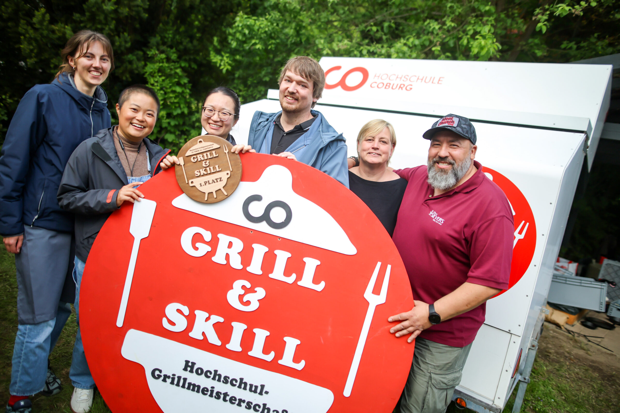 Sechs Personen lächeln und posieren im Freien mit einem großen roten "Grill & Skill"-Schild und einem hölzernen Preis der Hochschul-Grillmeisterschaft 2026. Hinter ihnen ist ein Anhänger der Hochschule Coburg zu sehen, im Hintergrund stehen Bäume. at Hochschule Coburg