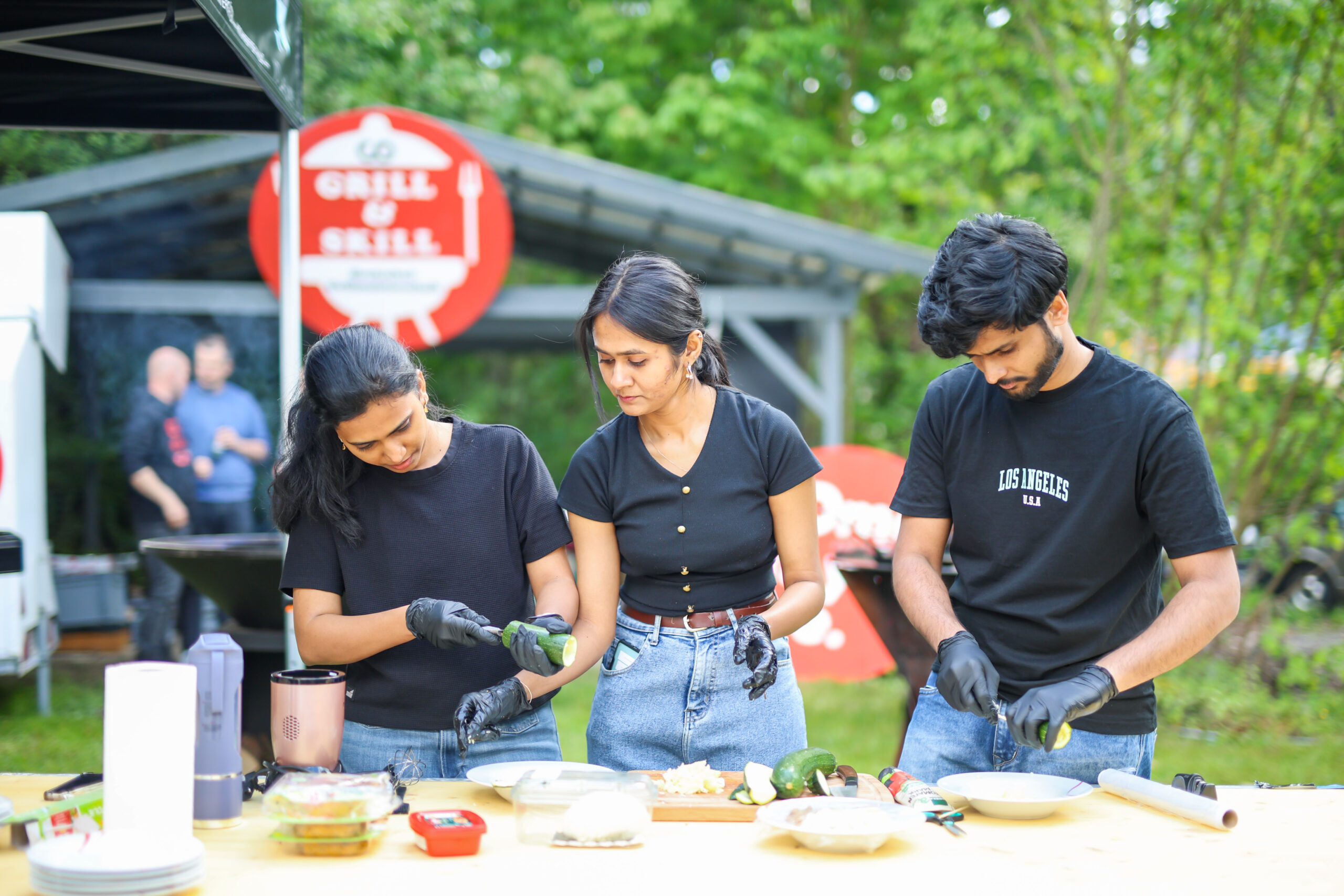 Drei Personen in schwarzen Hemden und Handschuhen bereiten an einem Tisch im Freien das Essen für die Hochschul-Grillmeisterschaft vor, umgeben von Gemüse wie Zucchini und Tofuschale. Im Hintergrund erscheinen ein "Grill & Chill"-Schild und Grünpflanzen. at Hochschule Coburg