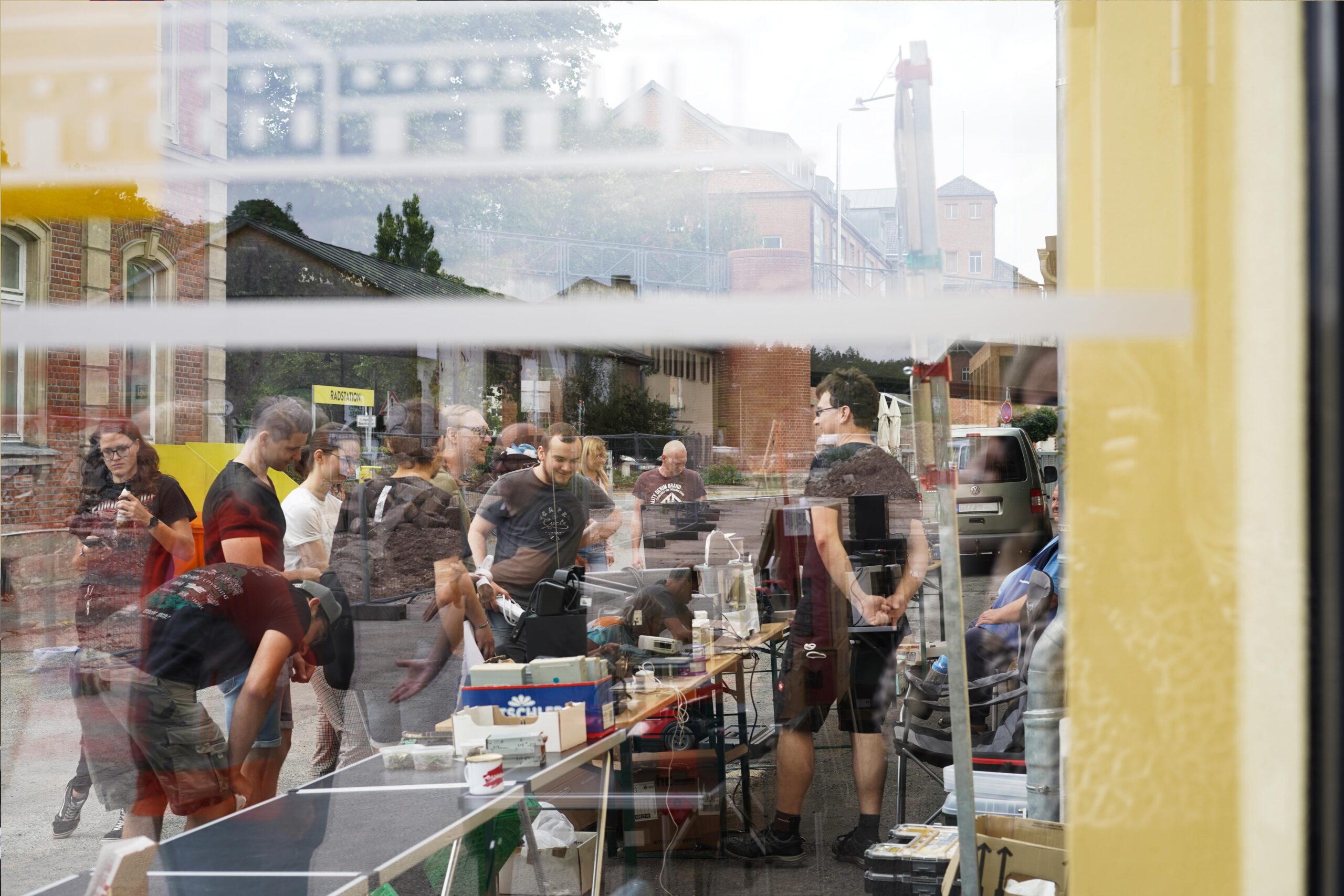 MAKE2026 Eine Gruppe von Menschen versammelt sich im Freien um Tische mit Elektronik und Werkzeugen, gesehen durch ein spiegelndes Fenster. Die Spiegelung zeigt Gebäude, Bäume und verschiedene Geräte, wodurch eine vielschichtige, geschäftige Szene entsteht. at Hochschule Coburg
